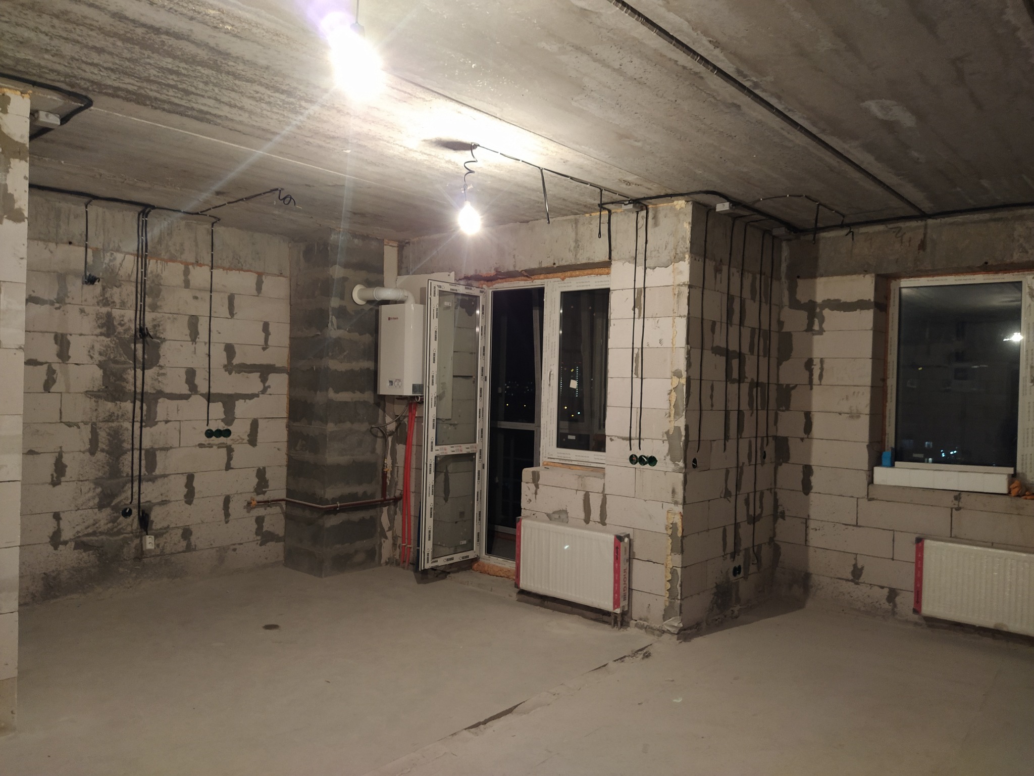 Surowy stan mieszkania w bloku: ściany z betonu komórkowego, instalacje elektryczne w peszlach, grzejnik pod oknem, widok na miasto nocą.