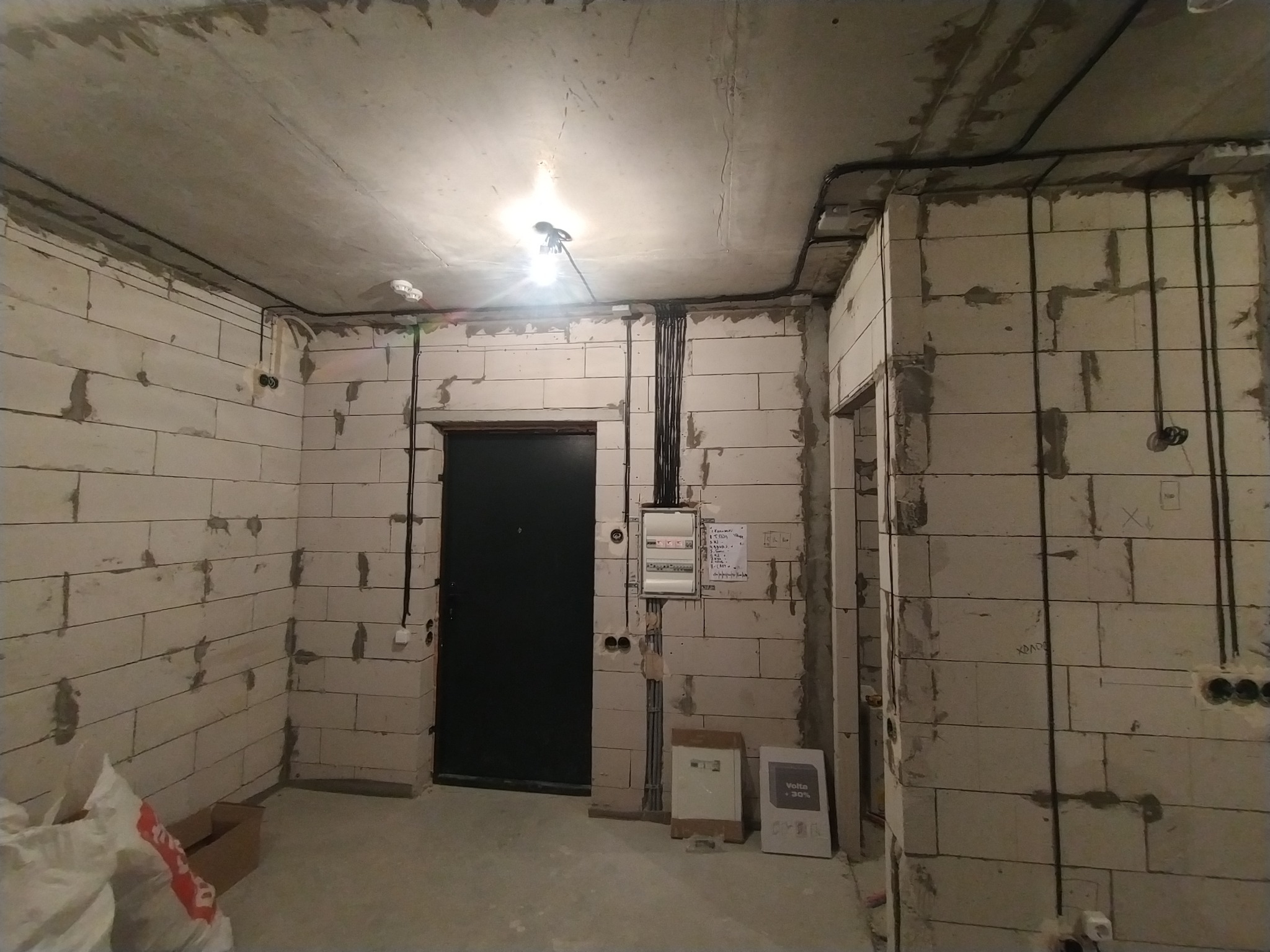 Surowy stan wnętrza z widoczną instalacją elektryczną: kable prowadzone po ścianach z betonu komórkowego, skrzynka rozdzielcza przy drzwiach, żarówka na kablu.
