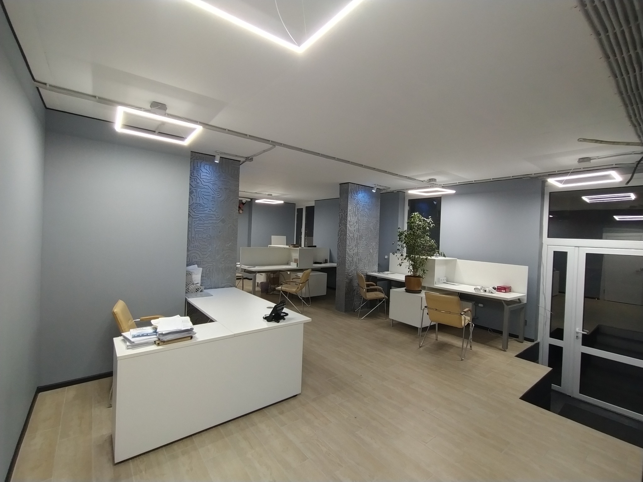 Nowoczesne biuro z białymi biurkami, krzesłami i oświetleniem LED. Ściany w odcieniach szarości z dekoracyjnymi panelami. Widok na przestrzeń biurową z rośliną doniczkową.