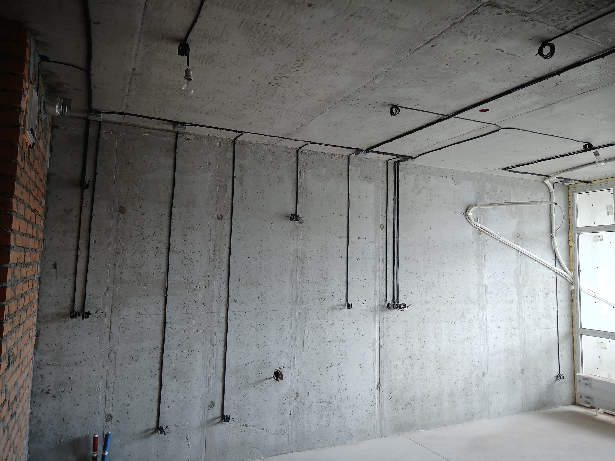 Surowy betonowy pokój z widoczną instalacją elektryczną: czarne kable prowadzone po suficie i ścianach, przygotowane do podłączenia. Widoczna żarówka na kablu.