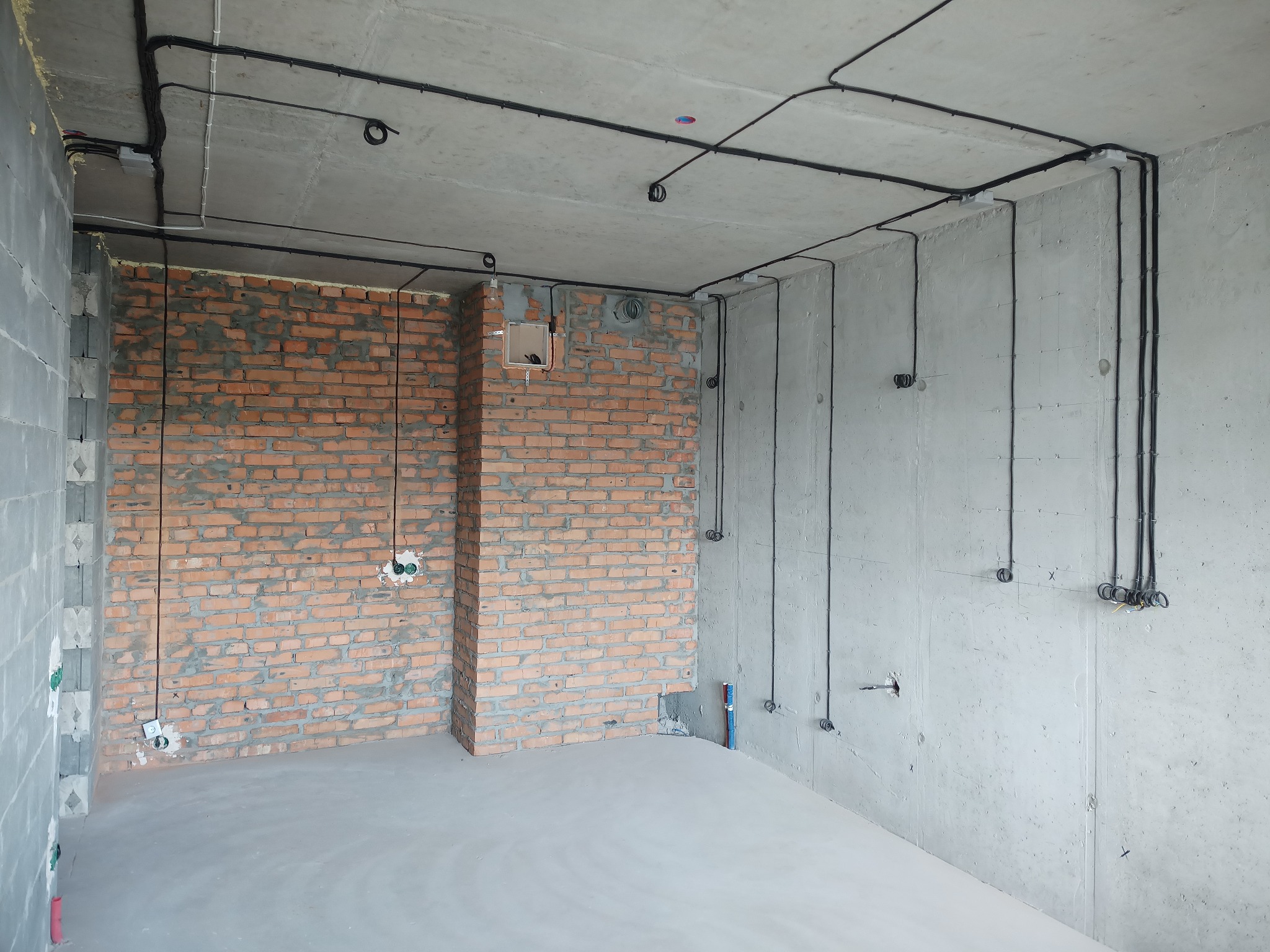 Surowy pokój z instalacją elektryczną: kable na suficie i ścianach, puszki pod gniazdka w ścianie z cegły i betonu. Wylewka na podłodze. Wnętrze w trakcie prac.