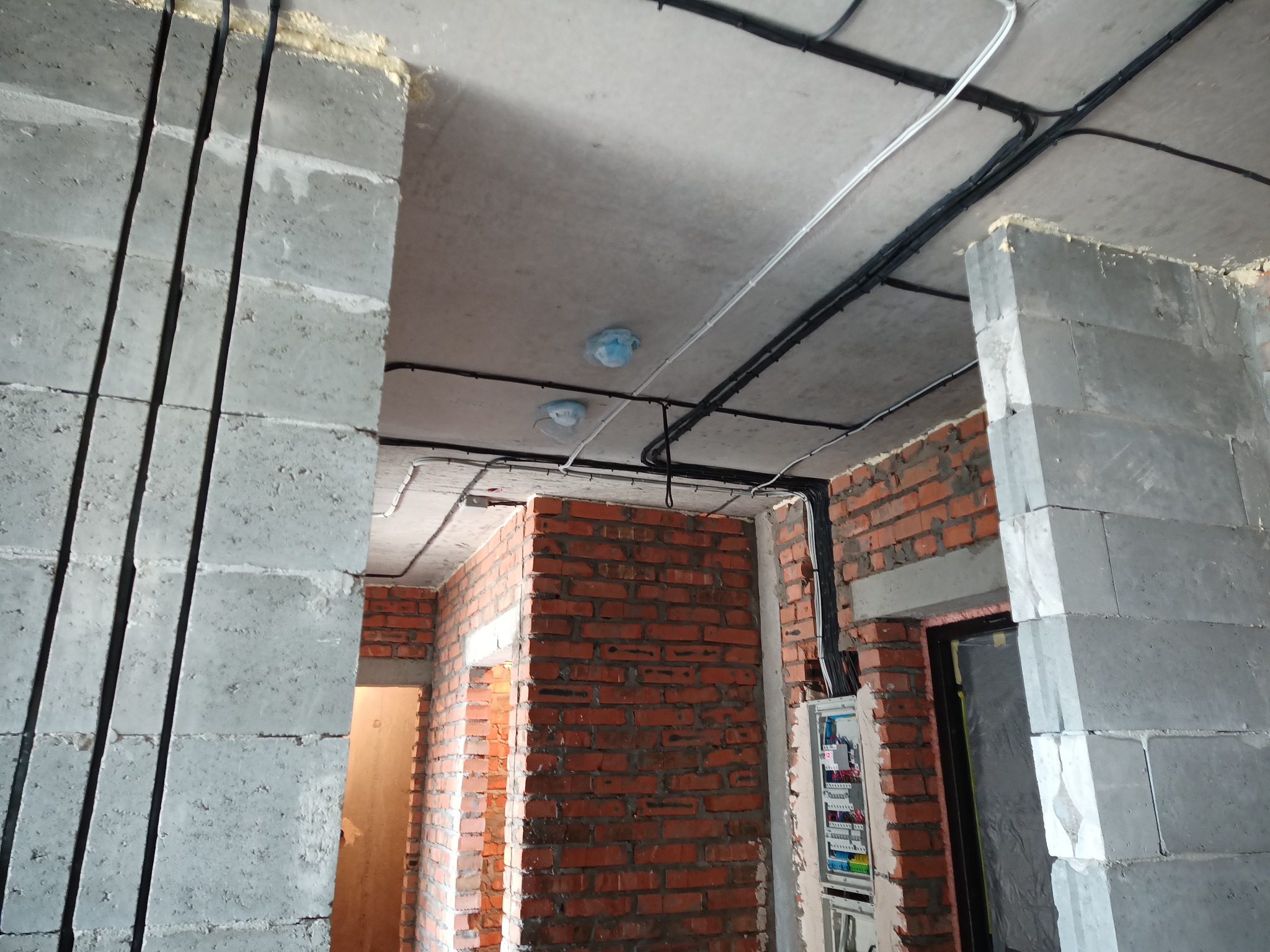 Wnętrze w trakcie prac: surowy beton, cegła, instalacje elektryczne w peszlach na suficie, skrzynka rozdzielcza. Widoczne zabezpieczenia na oświetleniu.