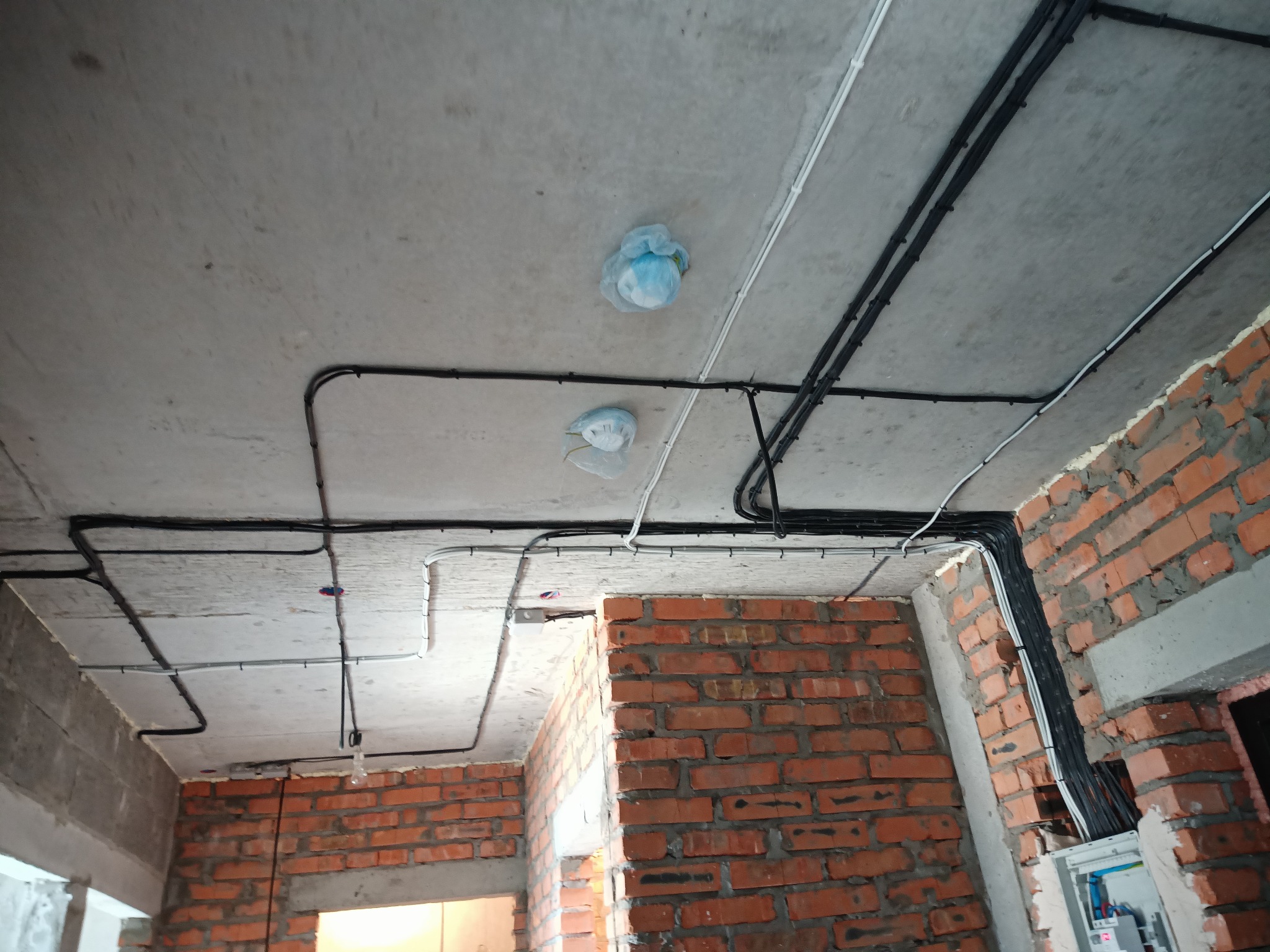 Surowy strop z instalacją elektryczną w peszlach, widoczne kable i puszki zabezpieczone folią. Ściany z czerwonej cegły w trakcie budowy. Rozdzielnia elektryczna.
