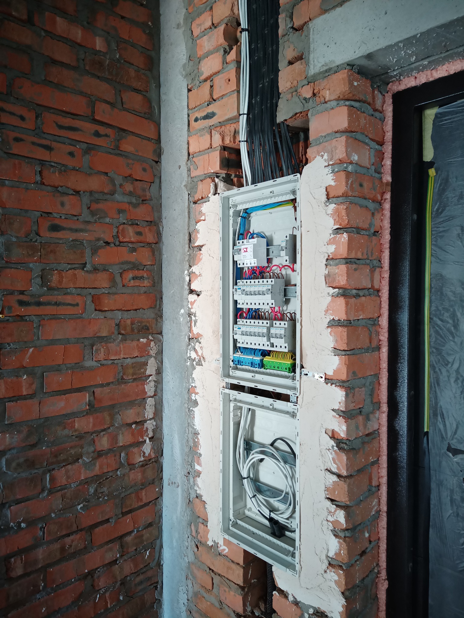 Rozdzielnica elektryczna w surowym stanie, montowana w ścianie z cegły w Rzeszowie. Widoczne kable i zabezpieczenia. Prace instalacyjne w trakcie realizacji.