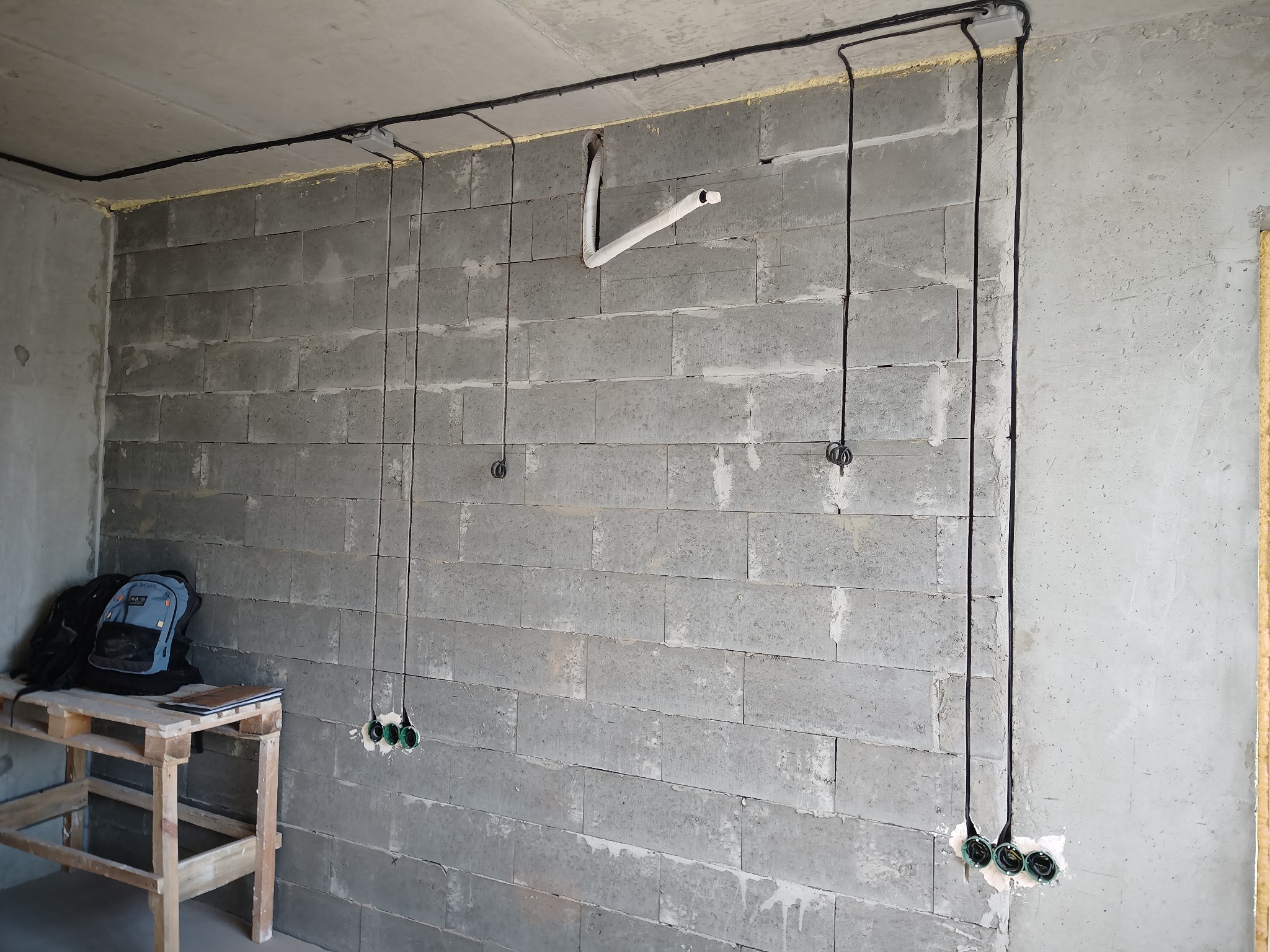 Surowa ściana z betonu komórkowego z poprowadzonymi przewodami elektrycznymi i puszkami pod gniazdka. Widoczny fragment instalacji wentylacyjnej i prowizoryczny stolik z palet.