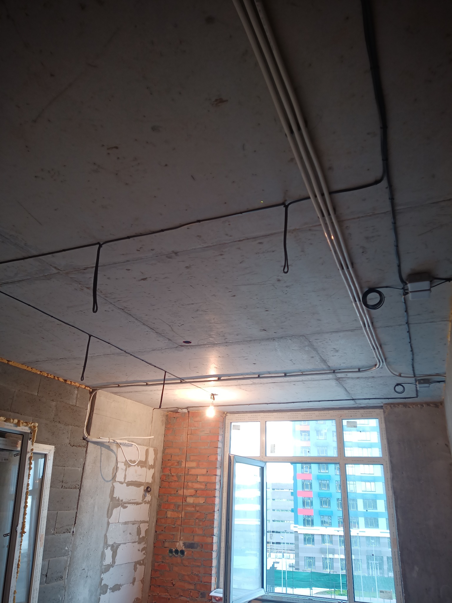 Surowy strop z widocznymi przewodami elektrycznymi i rurami instalacyjnymi w nowym mieszkaniu, okno z widokiem na budynek. Stan deweloperski.