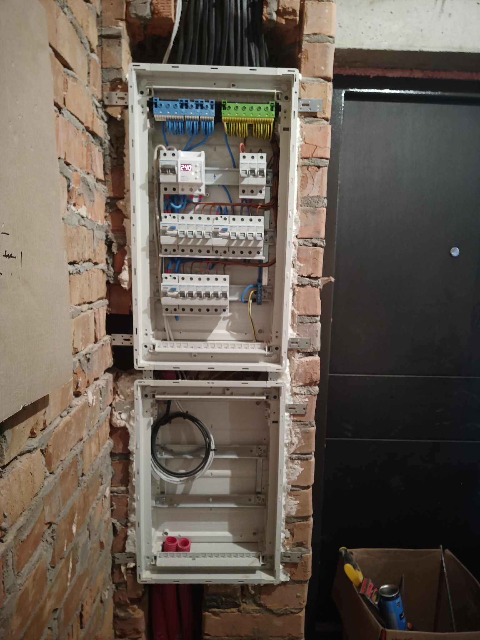 Otwarta skrzynka elektryczna z zainstalowanymi bezpiecznikami i przewodami, zamontowana na surowej, ceglanej ścianie wewnątrz budynku. Widoczne narzędzia w kartonie.