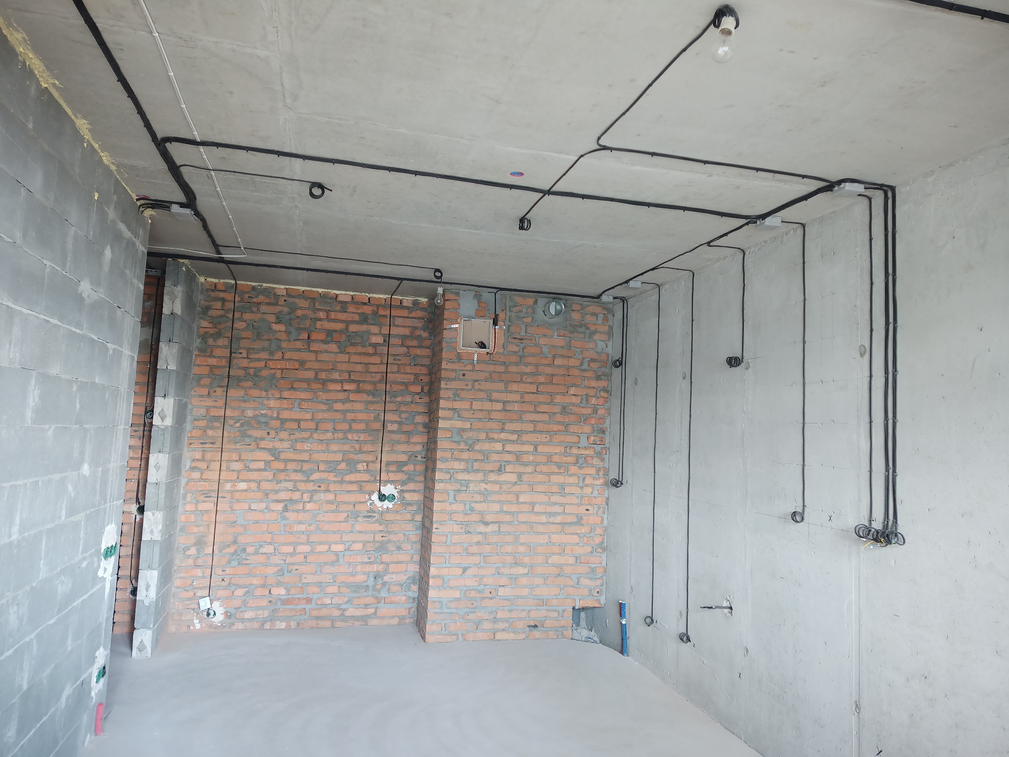 Surowy stan: czarne kable elektryczne prowadzone po suficie i ścianach w pomieszczeniu z surowymi ścianami z cegły i betonu. Widoczne puszki instalacyjne.