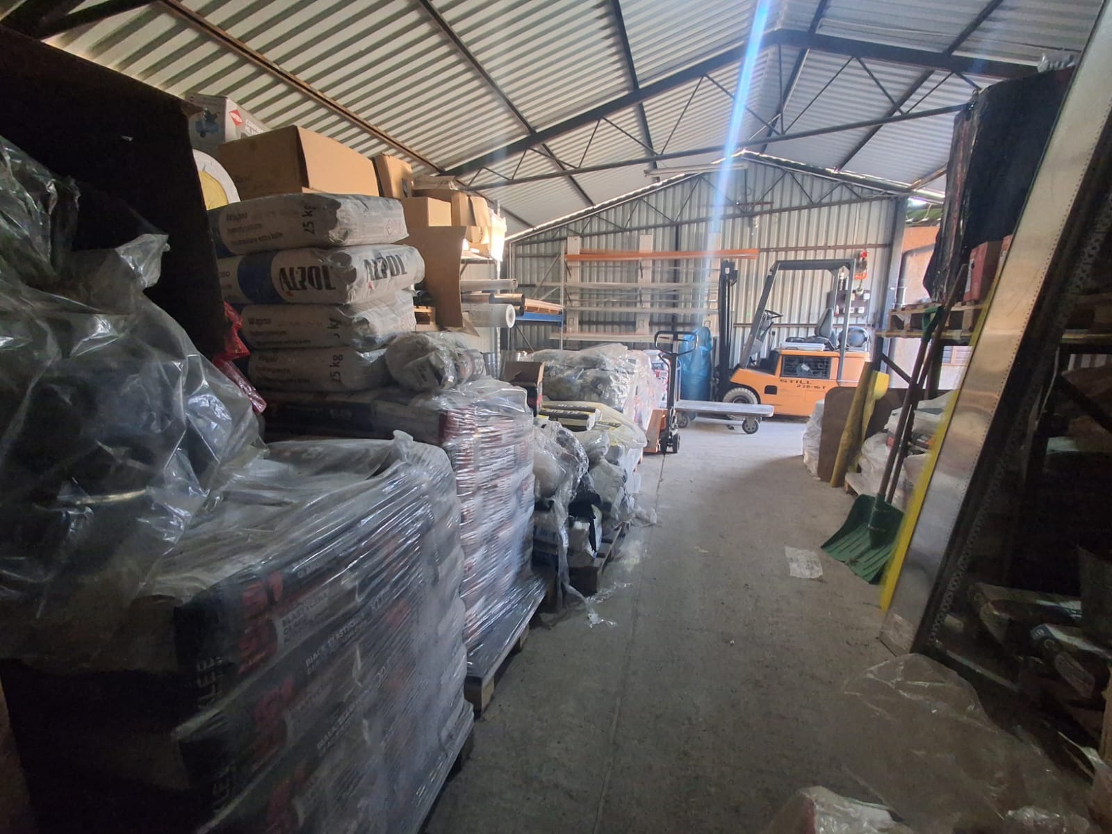 Wnętrze magazynu budowlanego z paletami materiałów pod folią, w tle wózek widłowy i regały. Metalowa konstrukcja dachu. Widok z perspektywy podłogi.