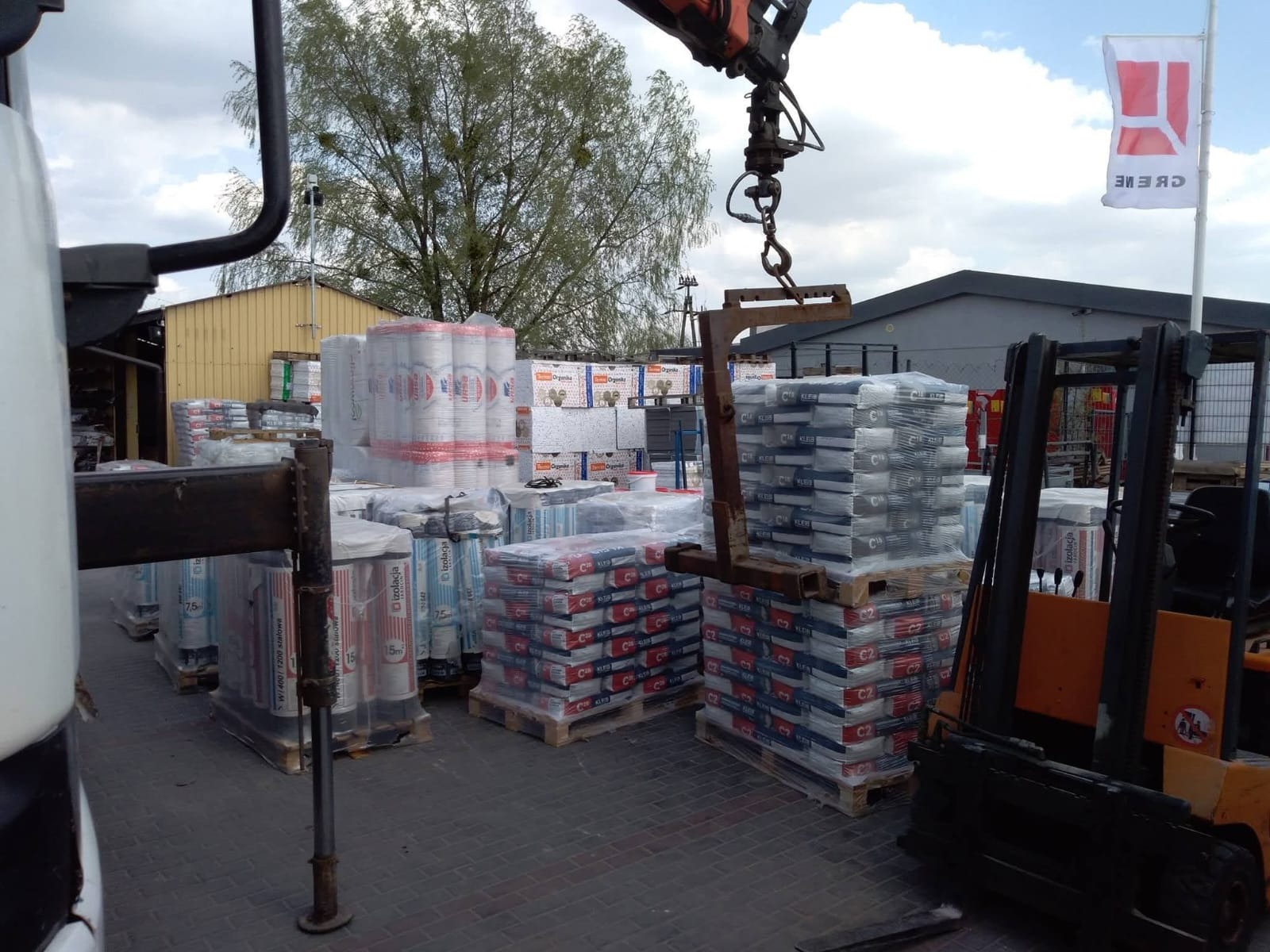 Skład budowlany: palety z workami kleju C2 i rolkami wełny mineralnej, transportowane wózkiem widłowym na tle magazynu i drzew. Widoczna flaga reklamowa.