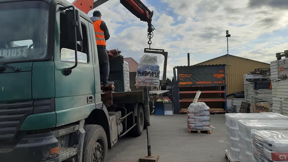 Rozładunek materiałów budowlanych za pomocą dźwigu z ciężarówki na placu składowym. Widoczny pracownik w kamizelce odblaskowej i palety z zapakowanymi produktami.