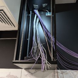 NEXTNET IT - Szafa rack z kablami sieciowymi w kolorach białym i fioletowym, widok z dołu, częściowo widoczna drabina. Instalacja sieci komputerowej w serwerowni.