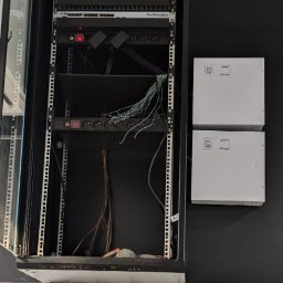 NEXTNET IT - Otwarta szafa rack z patch panelem, switchami i okablowaniem na tle czarnej ściany i marmurowej podłogi. Widoczne dwa białe panele z logo.