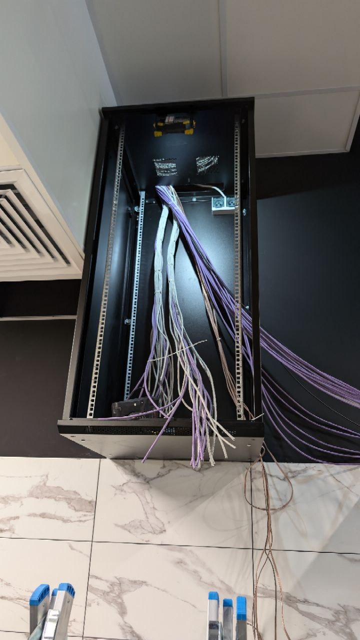 Szafa rack z kablami sieciowymi w kolorach białym i fioletowym, widok z dołu, częściowo widoczna drabina. Instalacja sieci komputerowej w serwerowni.
