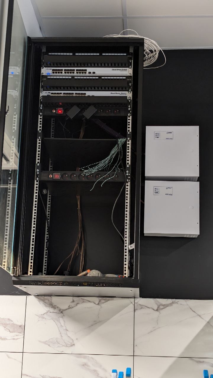 Otwarta szafa rack z patch panelem, switchami i okablowaniem na tle czarnej ściany i marmurowej podłogi. Widoczne dwa białe panele z logo.