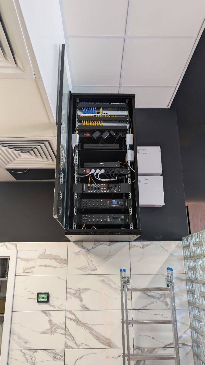 Zamontowana szafa rack z okablowaniem sieciowym i urządzeniami. Widoczna drabina i elementy wykończenia wnętrza. Instalacja w nowoczesnym biurze.