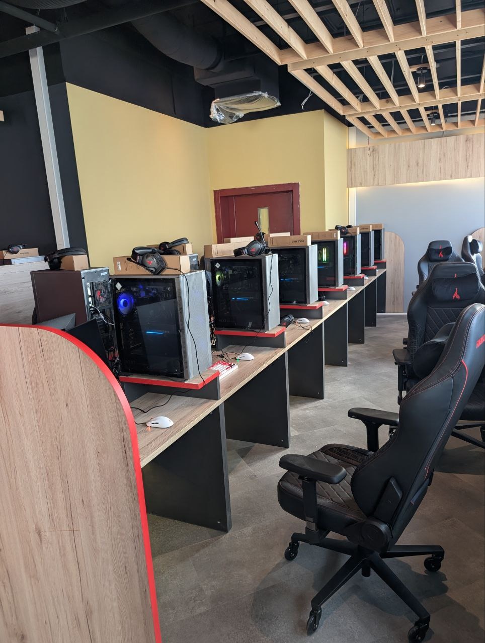 Sala gier z rzędem komputerów stacjonarnych, słuchawkami i ergonomicznymi fotelami. Widoczne drewniane biurka i sufit z odsłoniętą konstrukcją. Żółta ściana w tle.