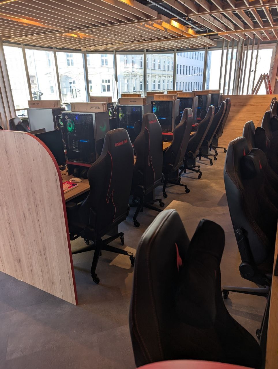 Sala z rzędem stanowisk komputerowych dla graczy, z czarnymi fotelami gamingowymi i oknami w tle. Widoczne komputery stacjonarne z podświetleniem RGB.