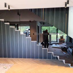 Bartinox balustrady szklane , nierdzewne - Nowoczesne schody z pionowymi panelami i szklaną balustradą, odbijającą wnętrze. Widoczny montażysta i elementy wykończeniowe na podłodze.