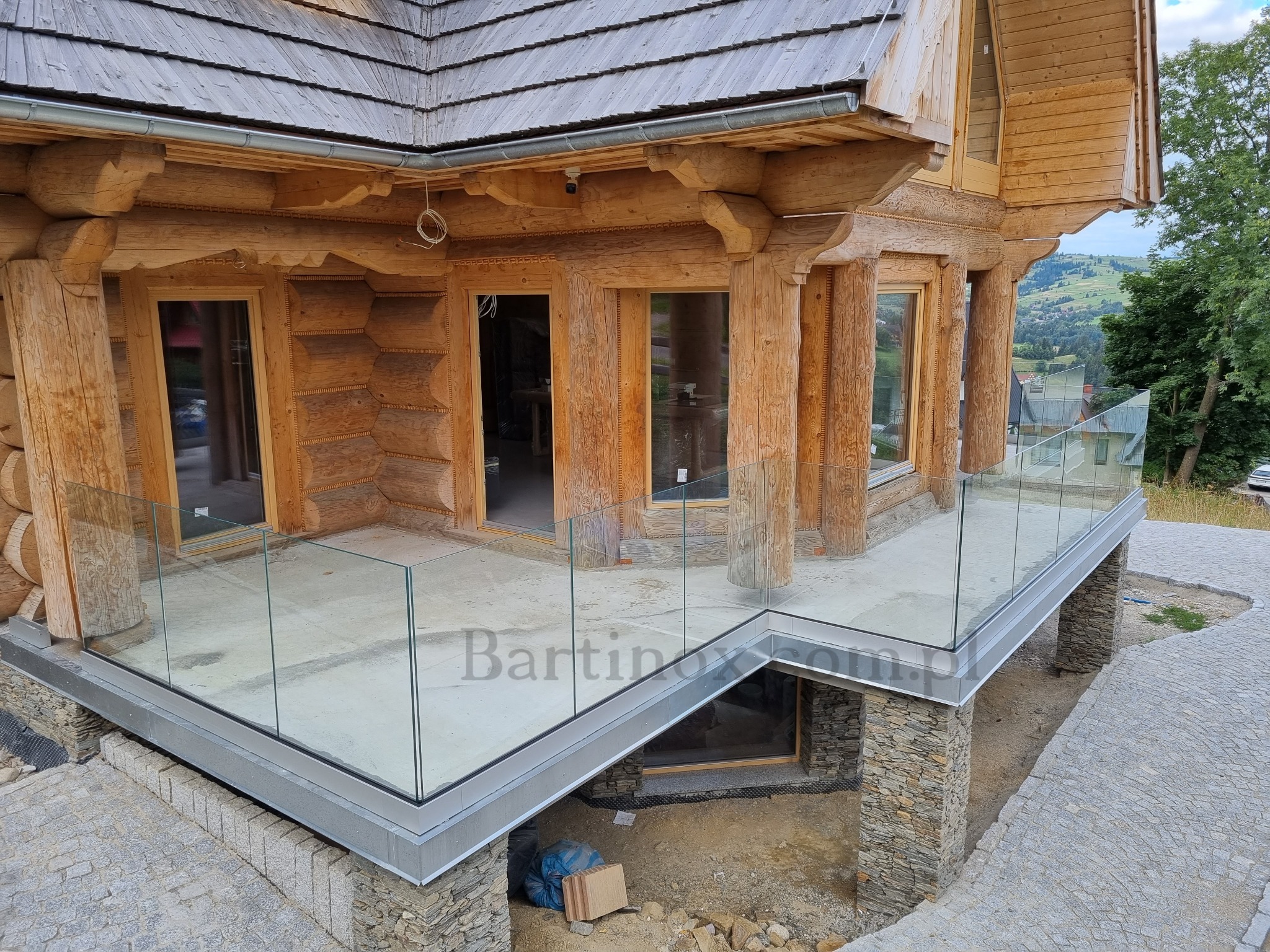 Szklana balustrada na tarasie drewnianego domu w stylu góralskim. Widok z zewnątrz, z elementami kamiennej podmurówki i górskiego krajobrazu w tle.