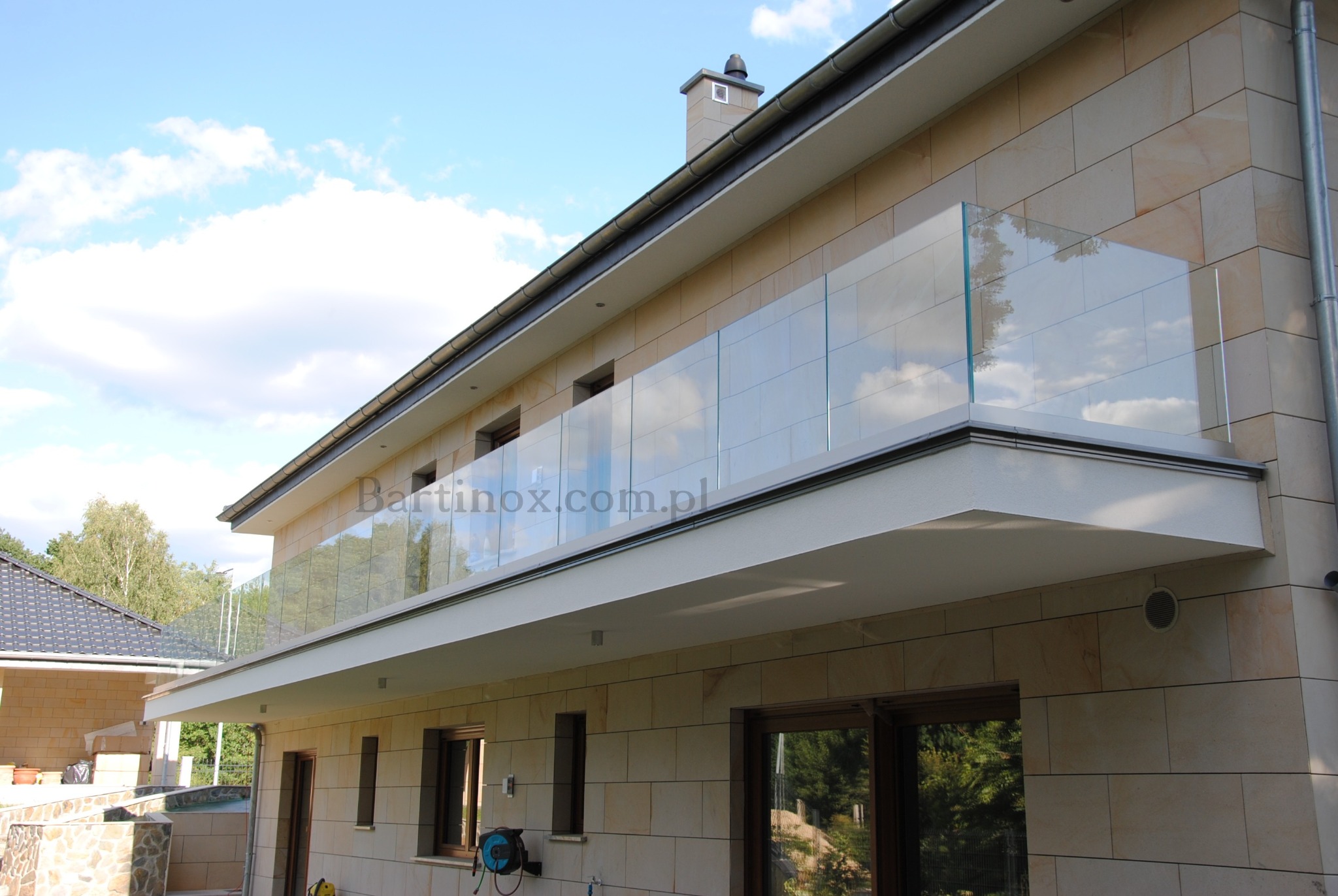 Nowoczesna, szklana balustrada balkonowa w domu z elewacją z piaskowca, widok z dołu. Przejrzyste szkło odbija błękitne niebo z chmurami.