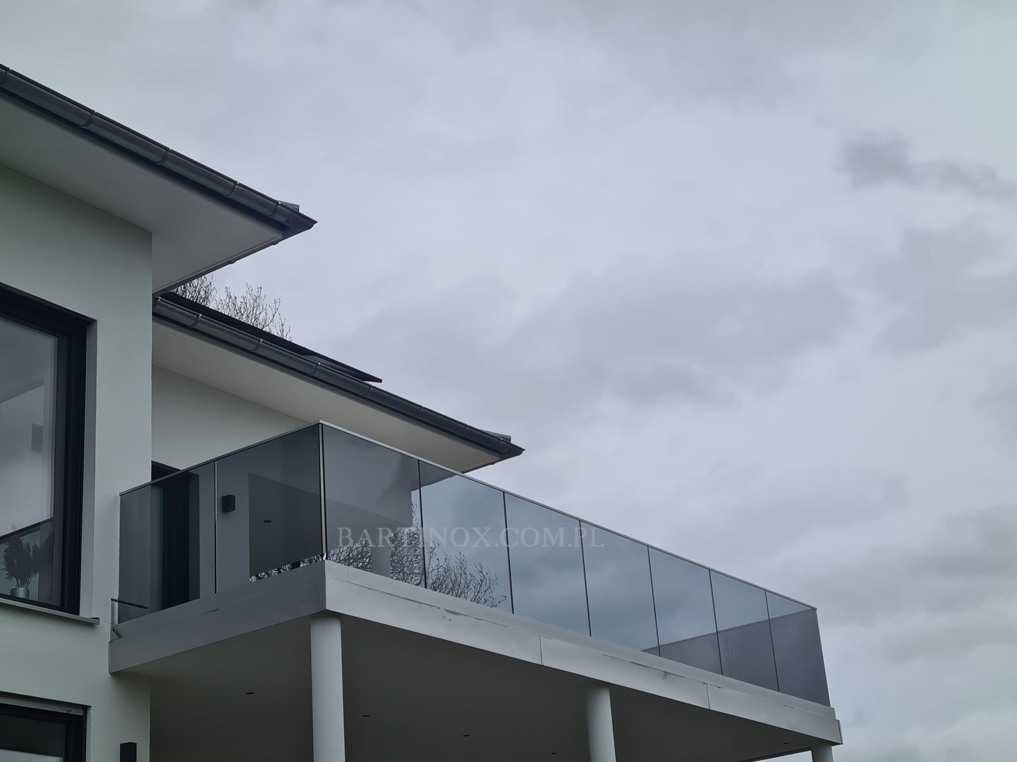 Nowoczesna balustrada szklana na balkonie domu jednorodzinnego, widok z dołu, pochmurne niebo w tle. Minimalistyczny design z kolumnami podtrzymującymi.