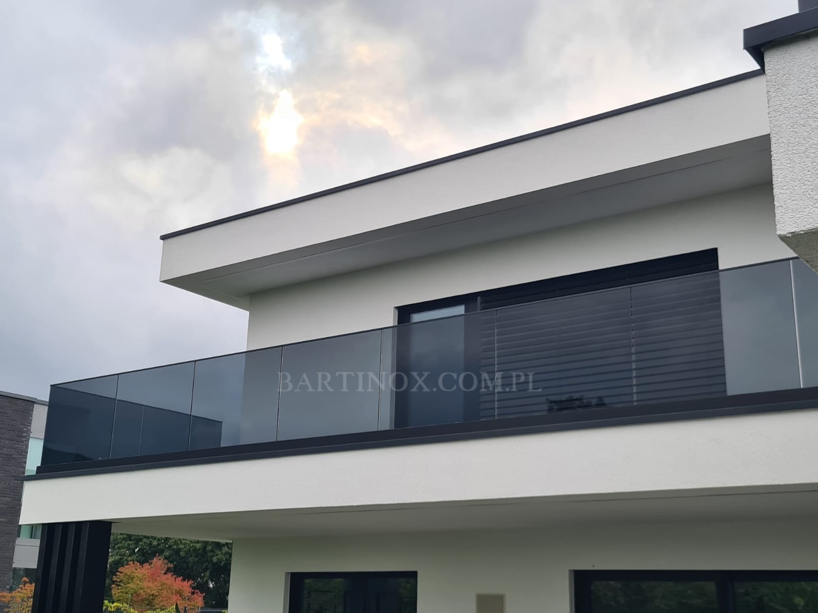 Nowoczesny balkon z szarą szklaną balustradą na tle pochmurnego nieba. Minimalistyczny design, widoczny adres strony internetowej na szkle. Elewacja budynku w jasnych barwach.