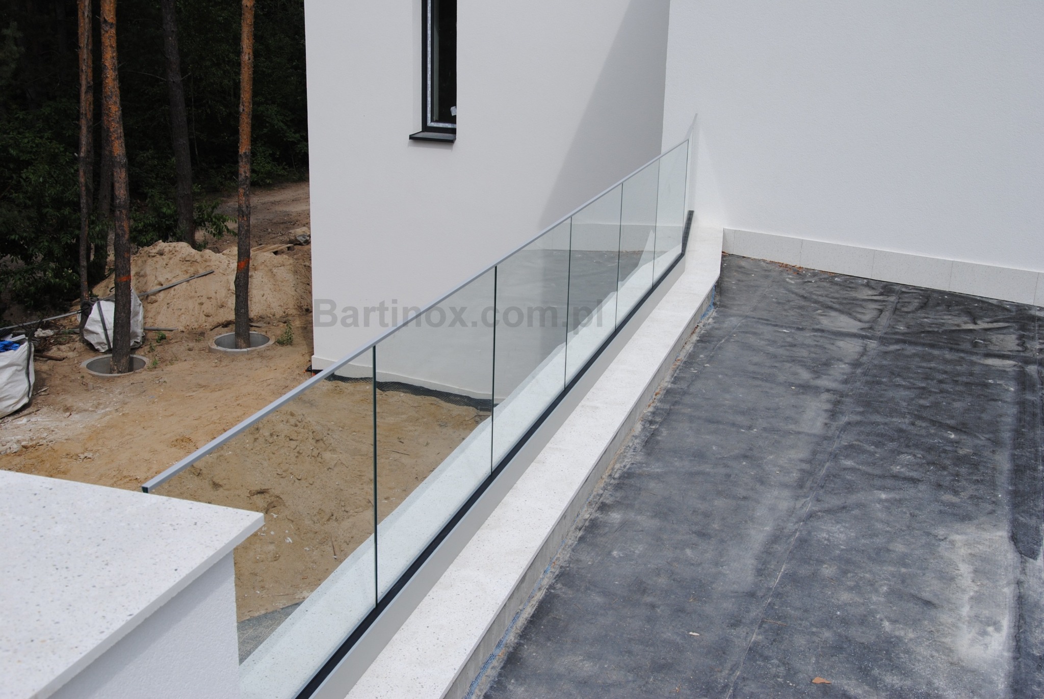 Nowoczesna, szklana balustrada z metalowymi słupkami na tarasie w trakcie wykańczania. Widok z góry, jasna kolorystyka, surowy stan otoczenia.