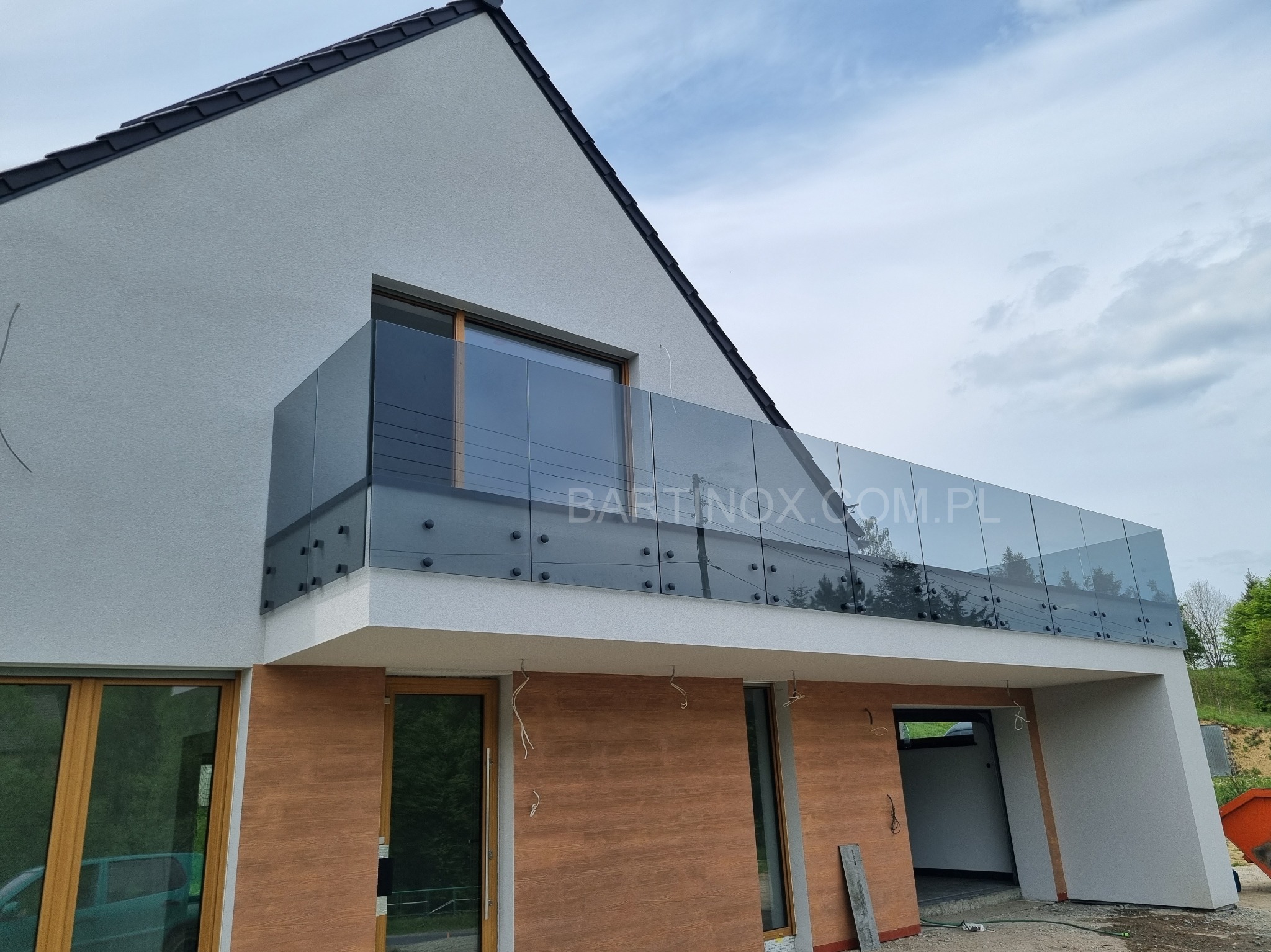 Nowoczesna balustrada szklana na balkonie domu jednorodzinnego, widok z dołu, z widocznym logo firmy na szkle. Minimalistyczny design, szare szkło, jasna elewacja.