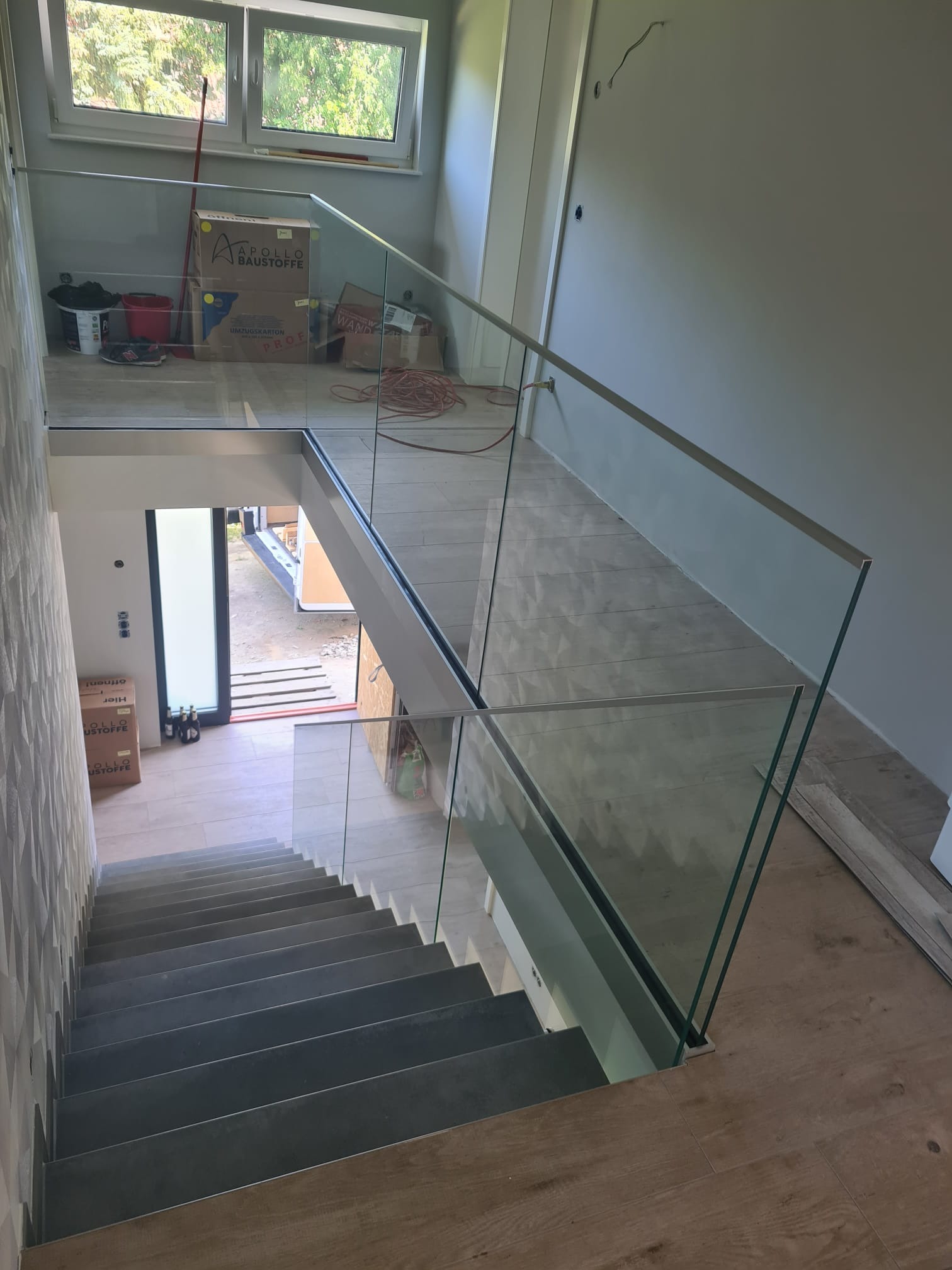Szklana balustrada z metalowymi elementami, zamontowana przy schodach wewnątrz budynku. Widok z góry, na pierwszym planie schody, w tle okno i kartony.