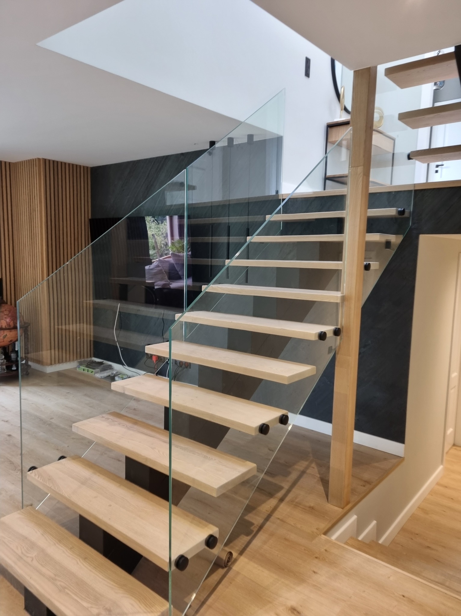 Nowoczesne schody z drewnianymi stopniami i szklaną balustradą, mocowaną na czarnych elementach. Jasne drewno kontrastuje z ciemną ścianą w tle.