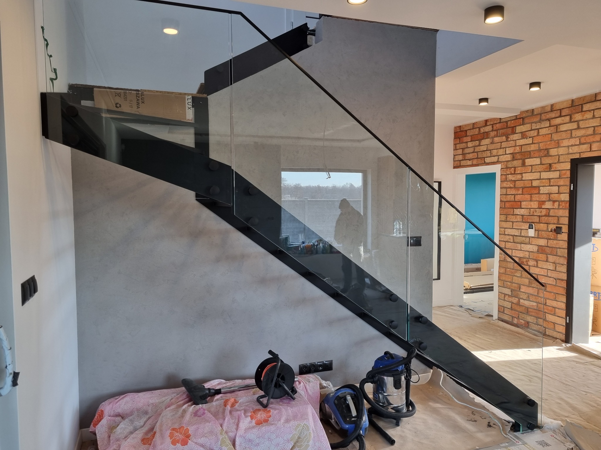 Szklana balustrada schodowa z czarnymi elementami mocującymi, widoczna w nowoczesnym wnętrzu z ceglaną ścianą i szarą, teksturowaną ścianą. W tle okno i osoba.