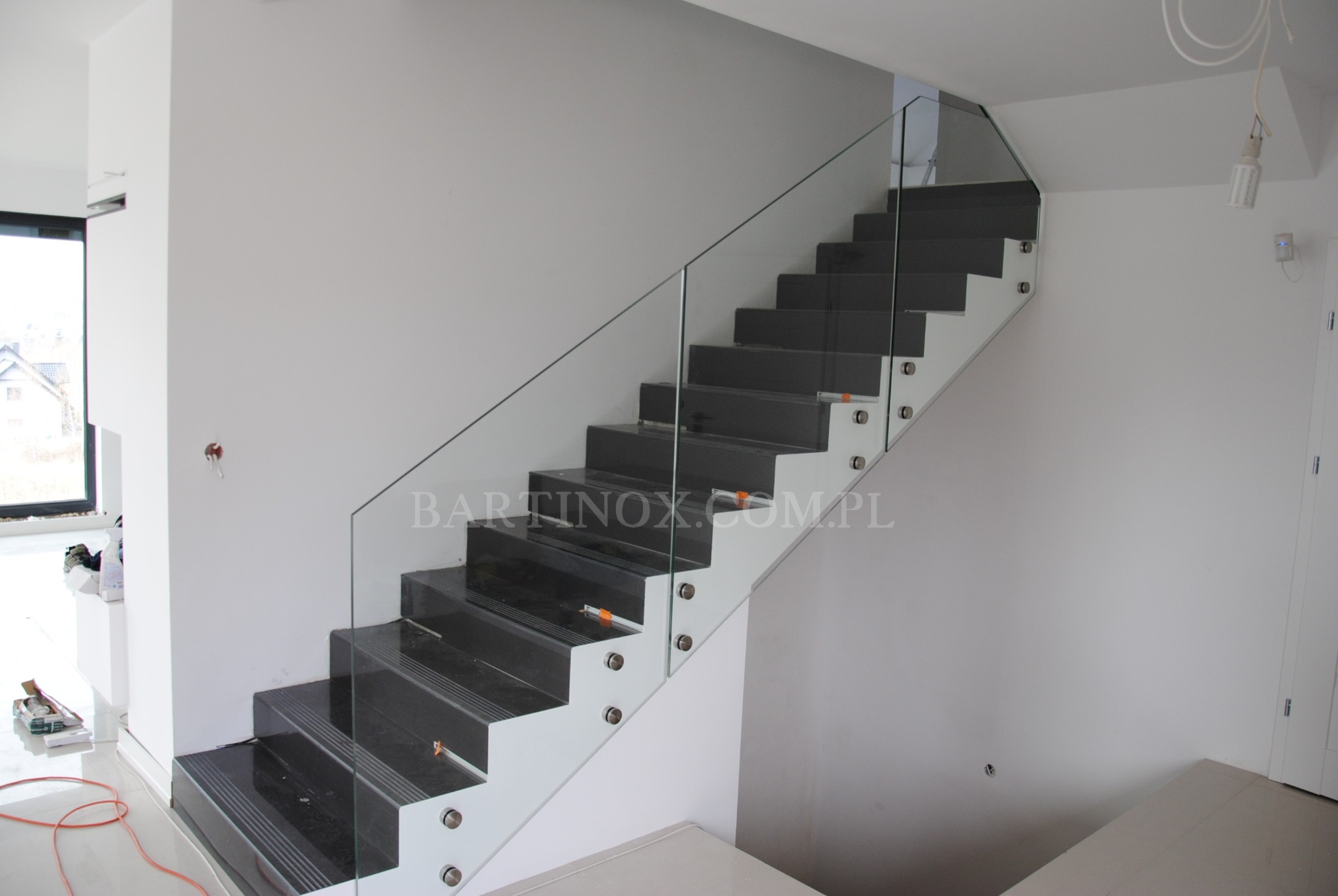 Nowoczesne schody z czarnymi stopniami i szklaną balustradą mocowaną na metalowych uchwytach. Minimalistyczny design w jasnym wnętrzu.