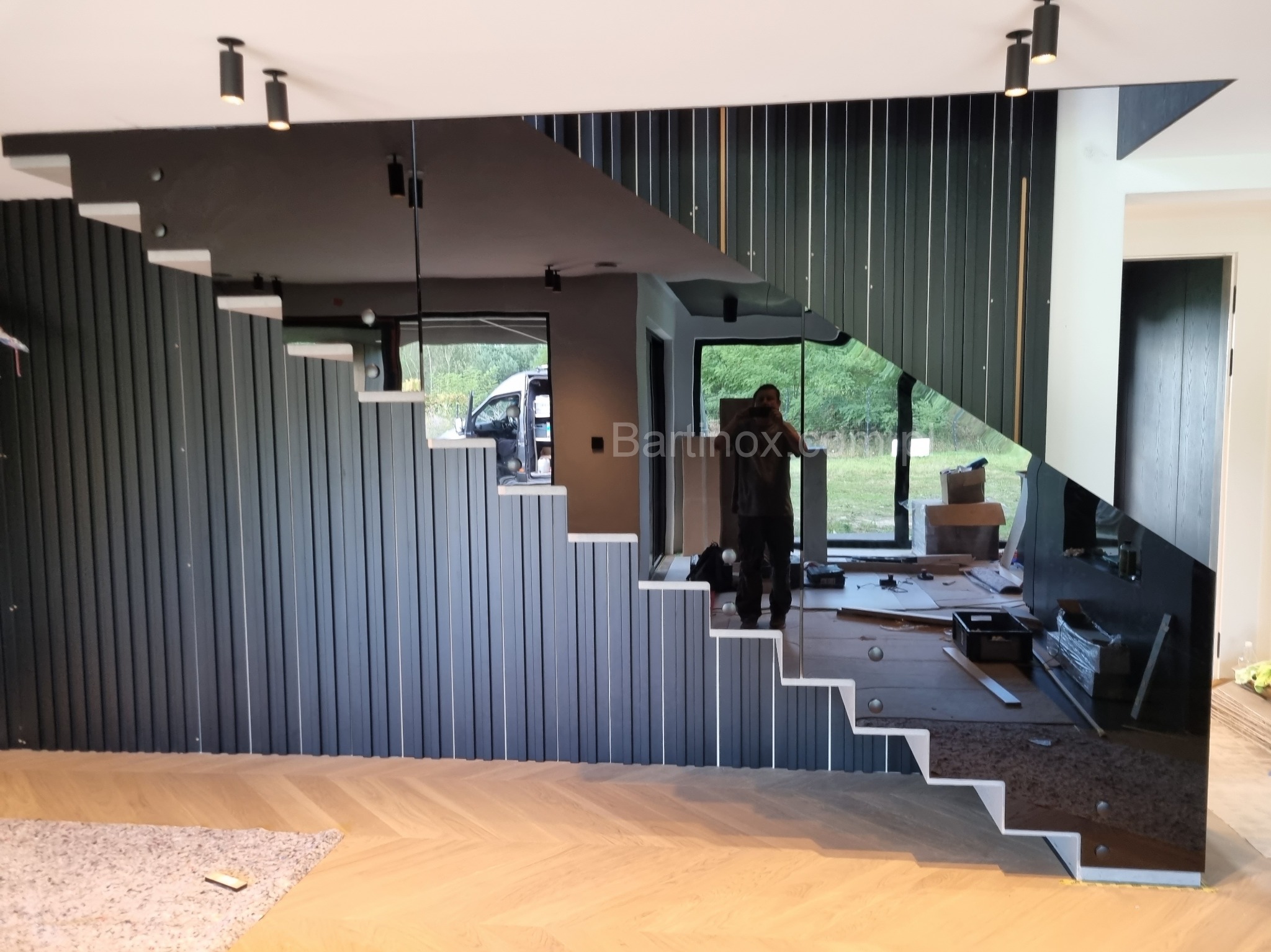 Nowoczesne schody z pionowymi panelami i szklaną balustradą, odbijającą wnętrze. Widoczny montażysta i elementy wykończeniowe na podłodze.