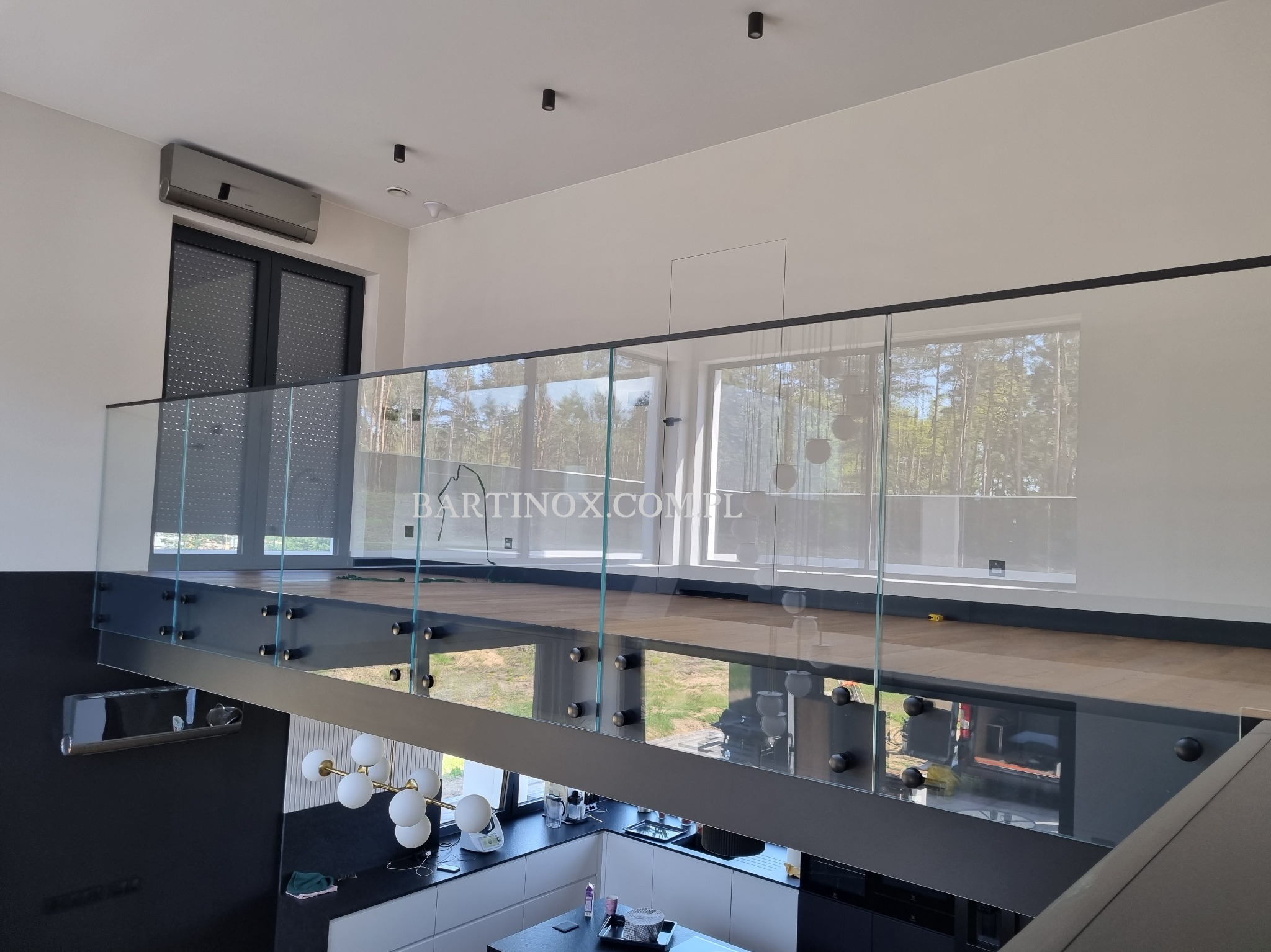 Nowoczesna balustrada szklana z czarnymi elementami mocującymi, widok z góry na kuchnię. Minimalistyczny design, jasne wnętrze z widokiem na zieleń.