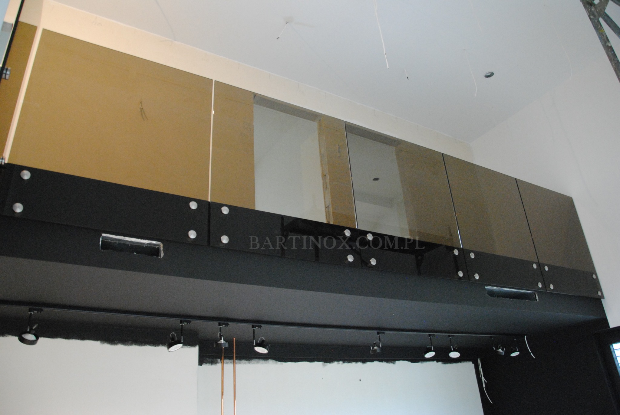 Szklana balustrada z czarnymi elementami mocującymi, widoczna od dołu, z oświetleniem punktowym. Na szkle widoczny adres strony internetowej.