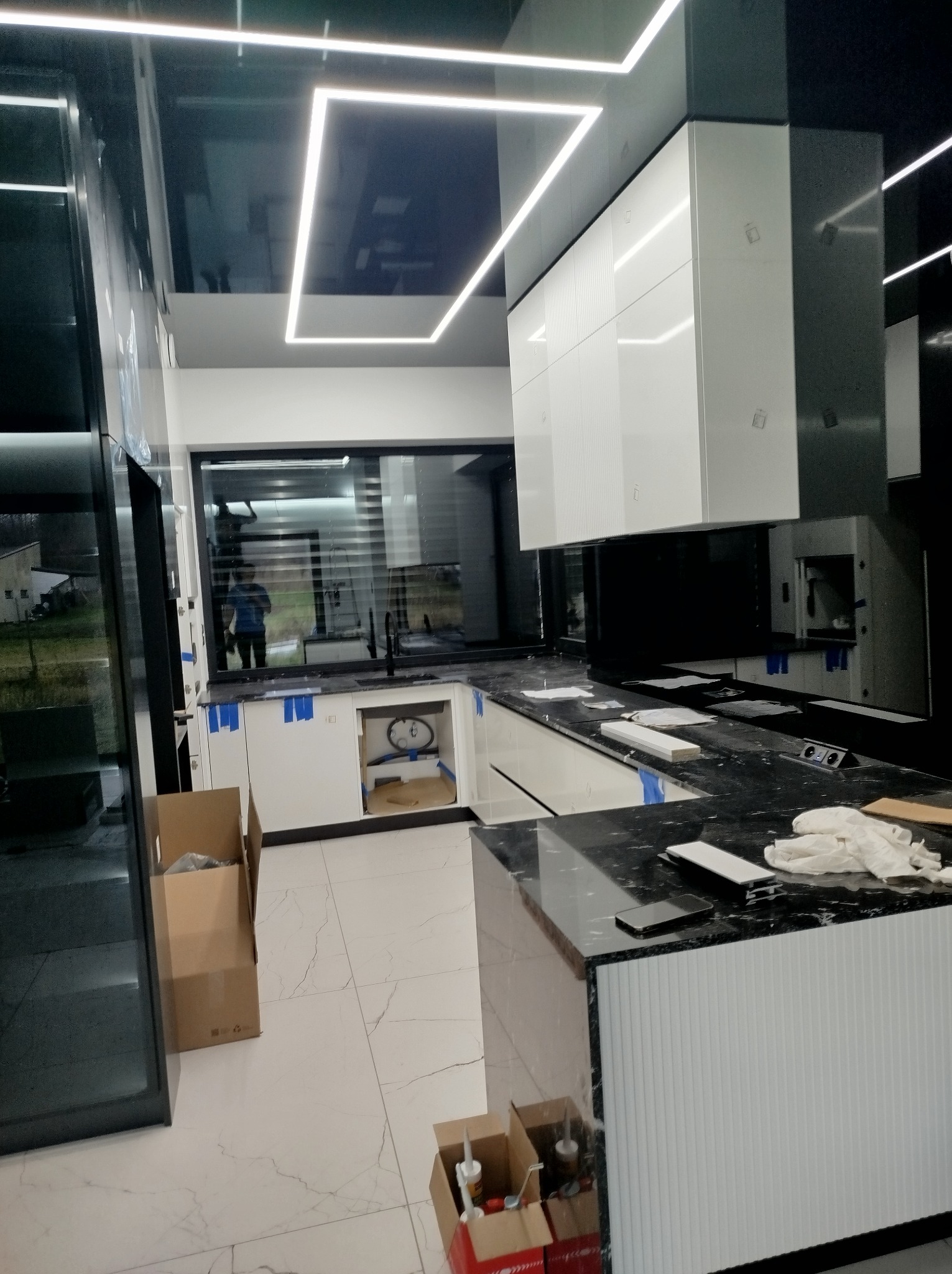 Nowoczesna kuchnia w trakcie montażu: białe szafki, czarny blat, marmurowe płytki podłogowe i linearne oświetlenie LED. Widoczne kartony i narzędzia.