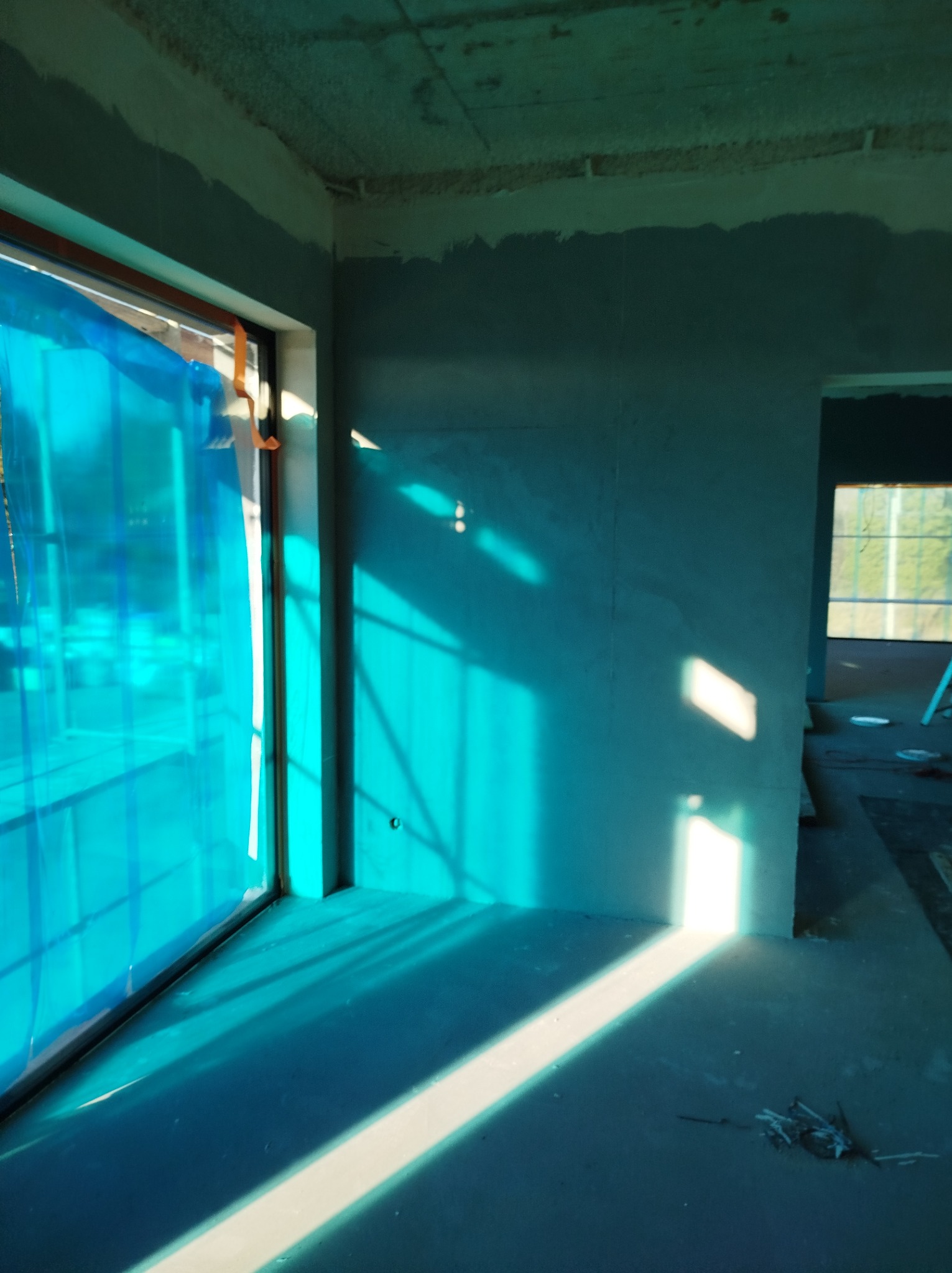 Wnętrze w trakcie szpachlowania ścian, z oknem zabezpieczonym folią i wyraźnymi promieniami słonecznymi. Widoczny surowy betonowy strop.