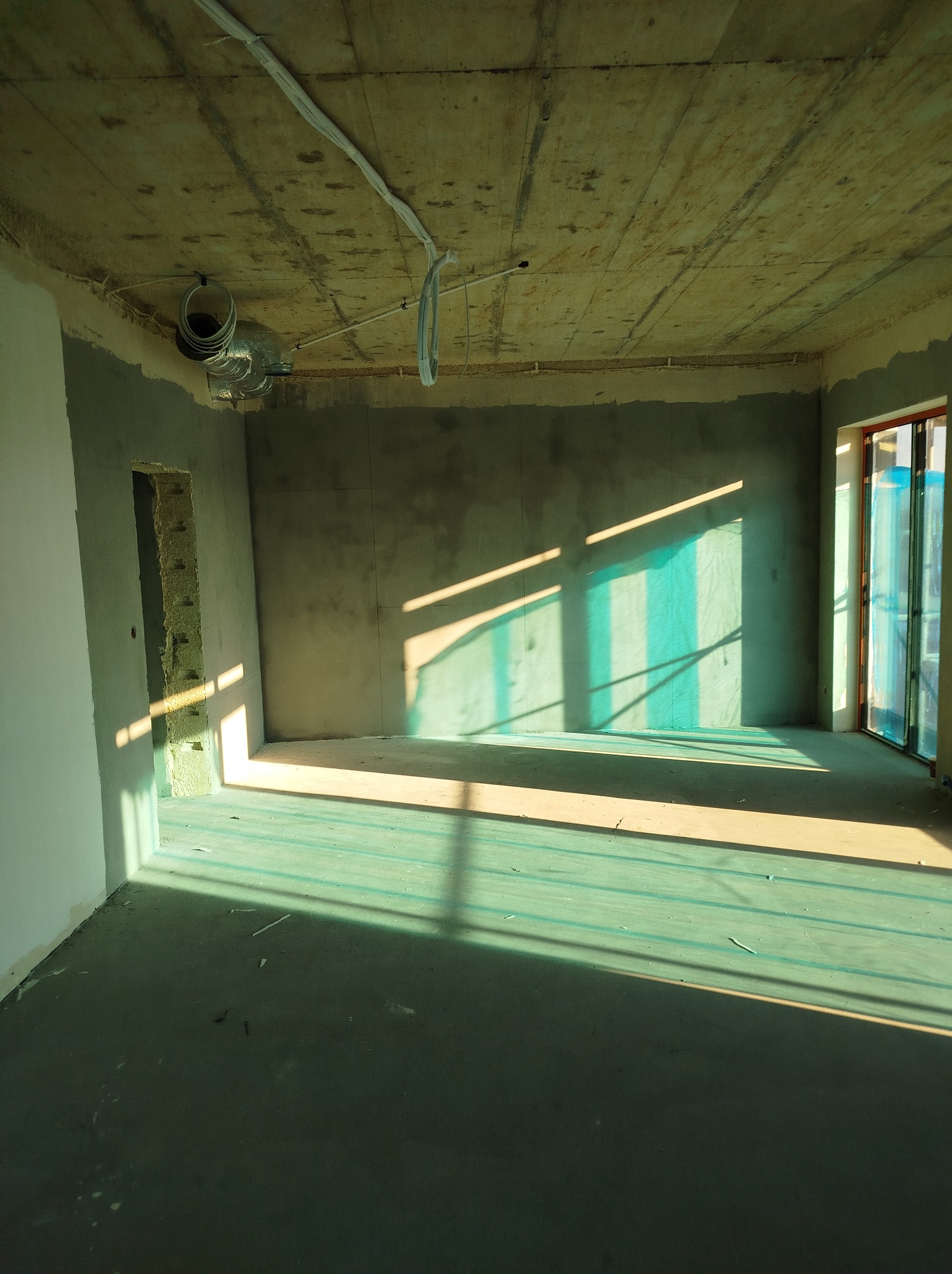 Surowe wnętrze w trakcie remontu: szpachlowane ściany, betonowy sufit z instalacjami, okno z folią ochronną, promienie słoneczne tworzą geometryczne cienie na podłodze.