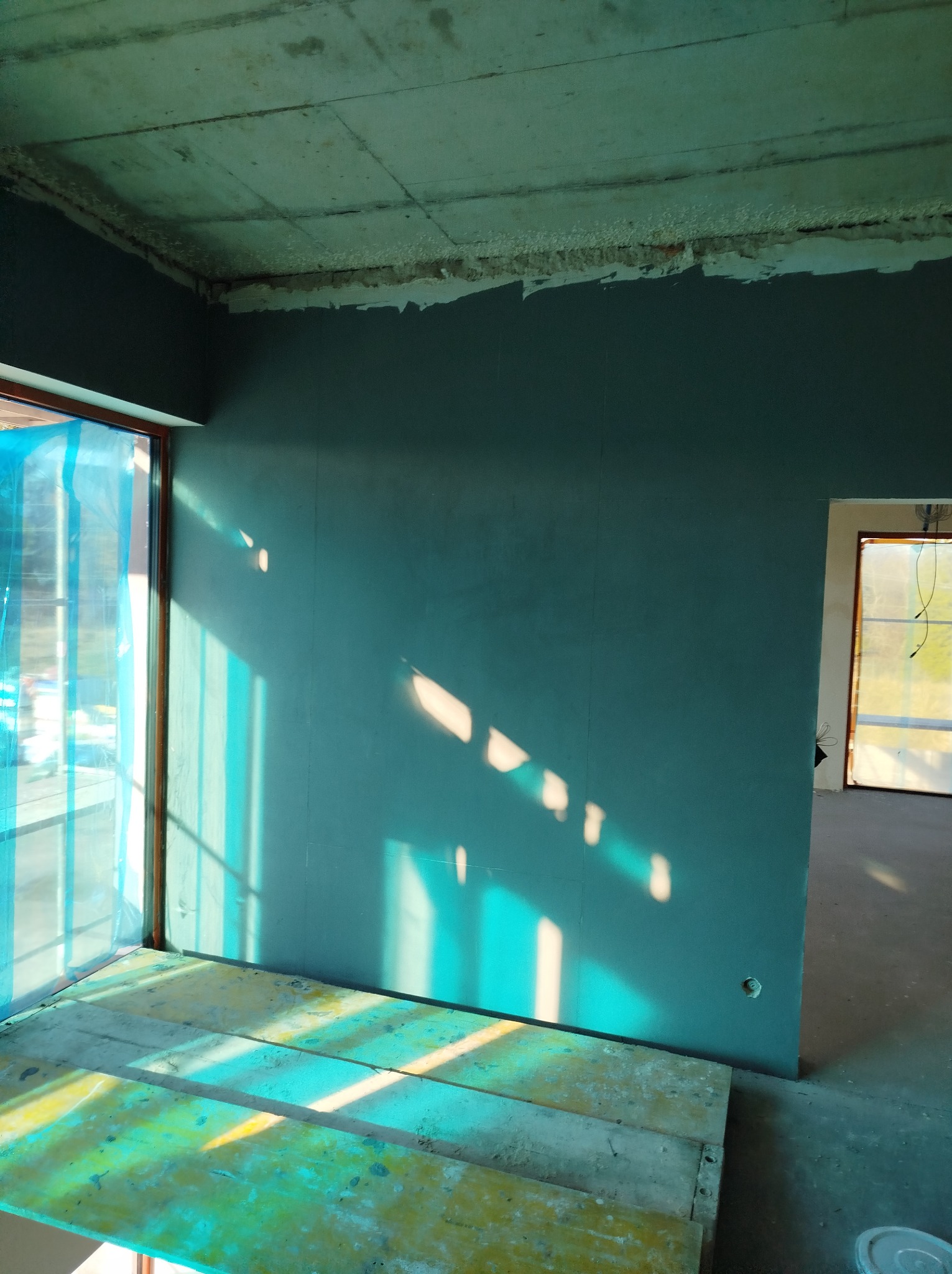 Ściana po szpachlowaniu w odcieniach szarości, z widocznymi promieniami słońca. W tle surowy betonowy sufit i okno zabezpieczone folią. Widoczny fragment rusztowania.