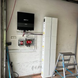 PHU Vika Kitaieva Most Energy - Montaż magazynu energii Encor i inwertera na ścianie garażu. Widoczne kable, skrzynki elektryczne i drabina. Podłoga z drobnymi zabrudzeniami.