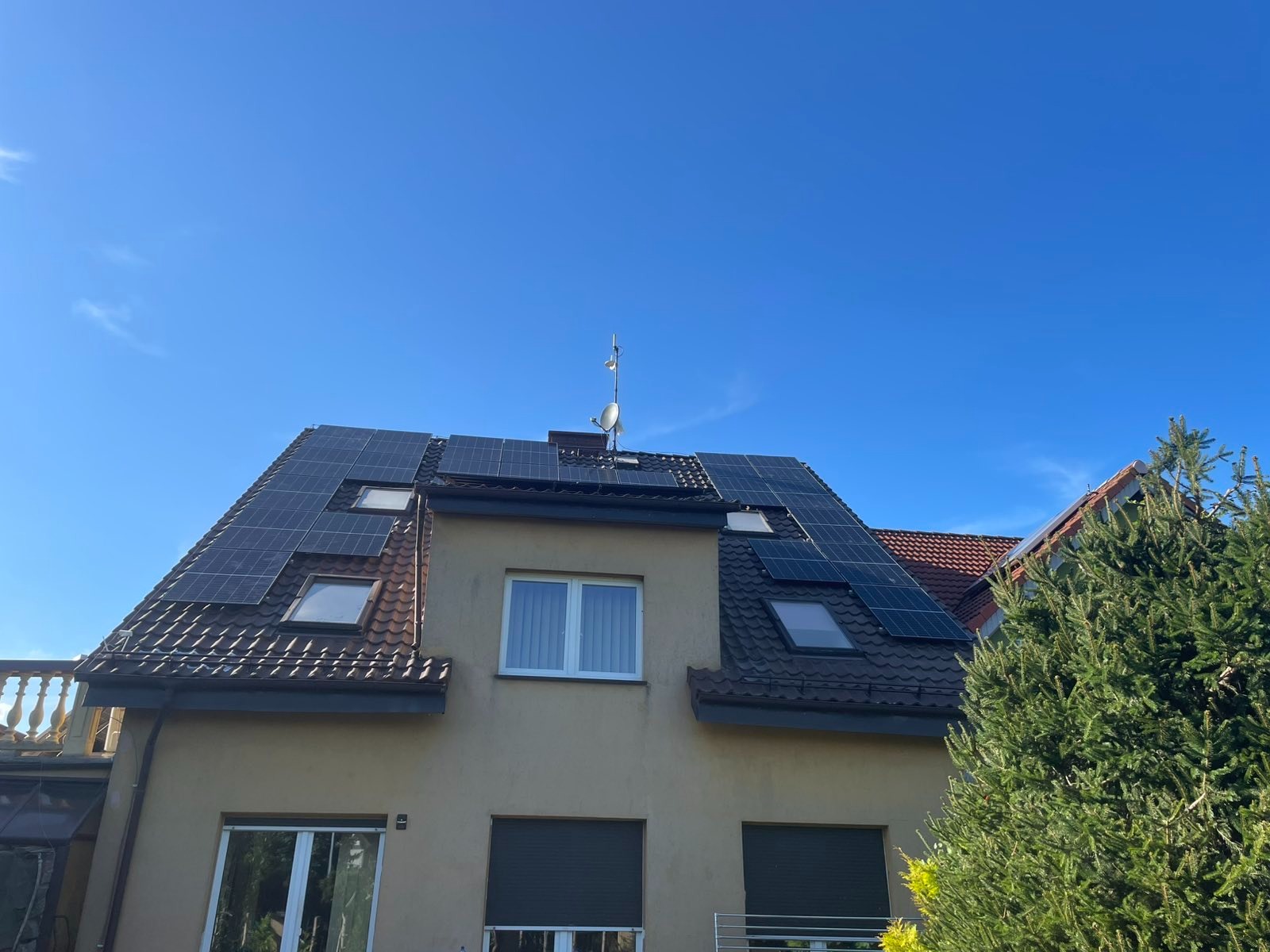 Dom z panelami słonecznymi na dachu z dachówką, okna dachowe, antena satelitarna, błękitne niebo. Widok z dołu, zieleń drzewa po prawej.