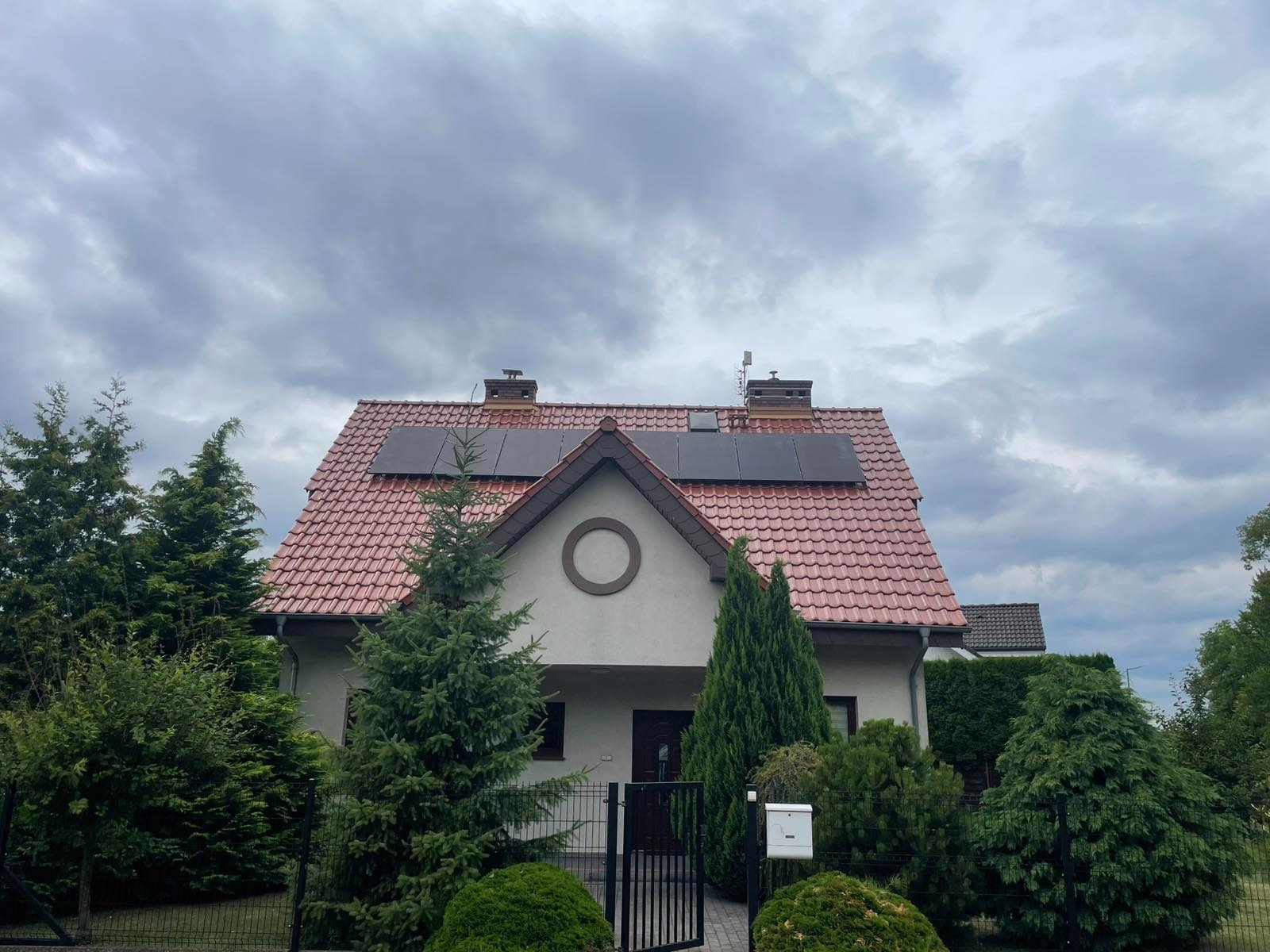 Dom jednorodzinny z panelami słonecznymi na dachu pokrytym czerwoną dachówką. Widok z poziomu gruntu, pochmurne niebo w tle. Ogród z iglakami.