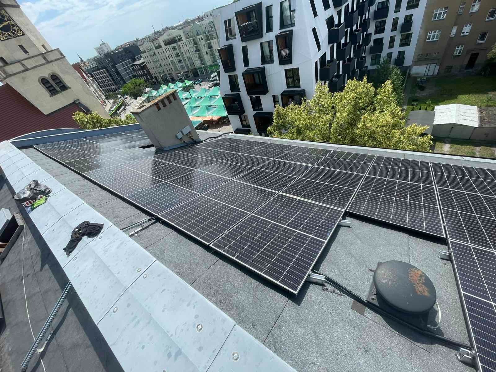 Panele słoneczne na dachu budynku w Poznaniu, z kominem i fragmentami zabudowy miejskiej w tle. Widok z góry, oświetlenie dzienne, instalacja fotowoltaiczna.