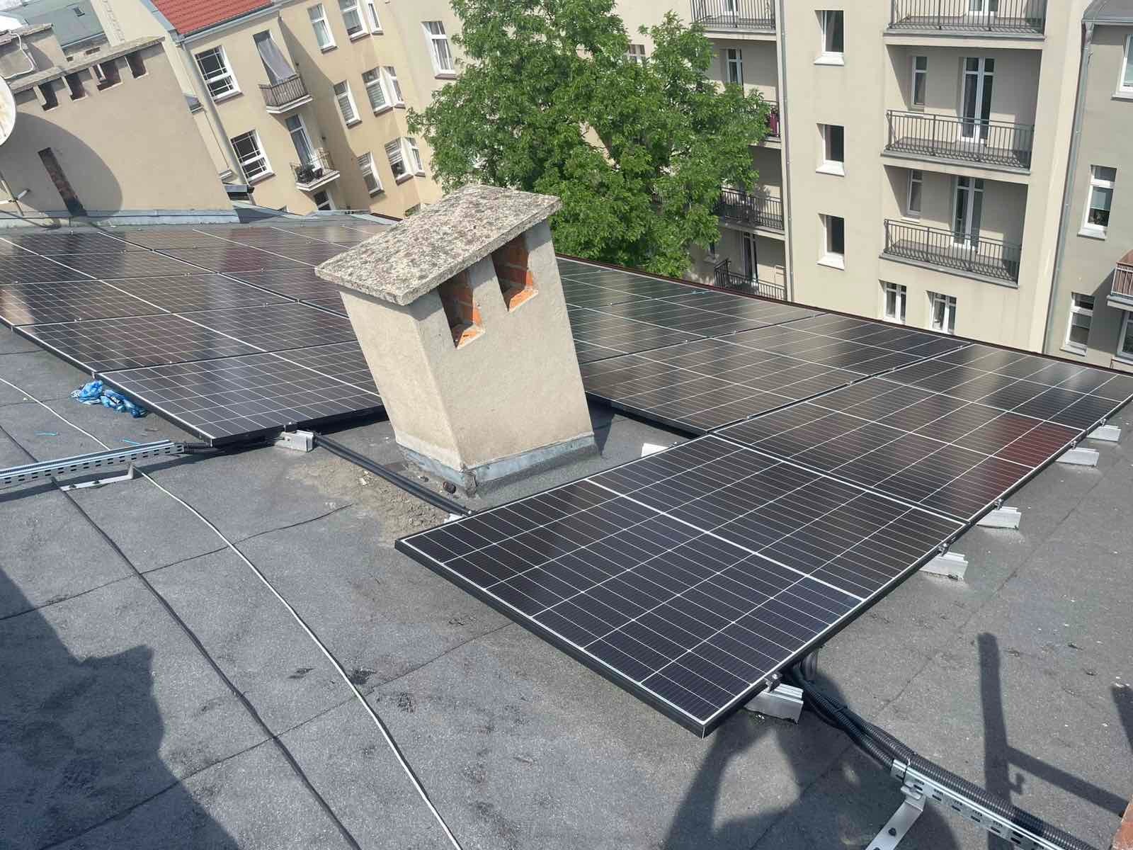 Panele słoneczne na dachu budynku mieszkalnego, z kominem w tle. Widoczne elementy montażowe i okablowanie. Ujęcie z perspektywy dachu, w tle budynki i drzewa.