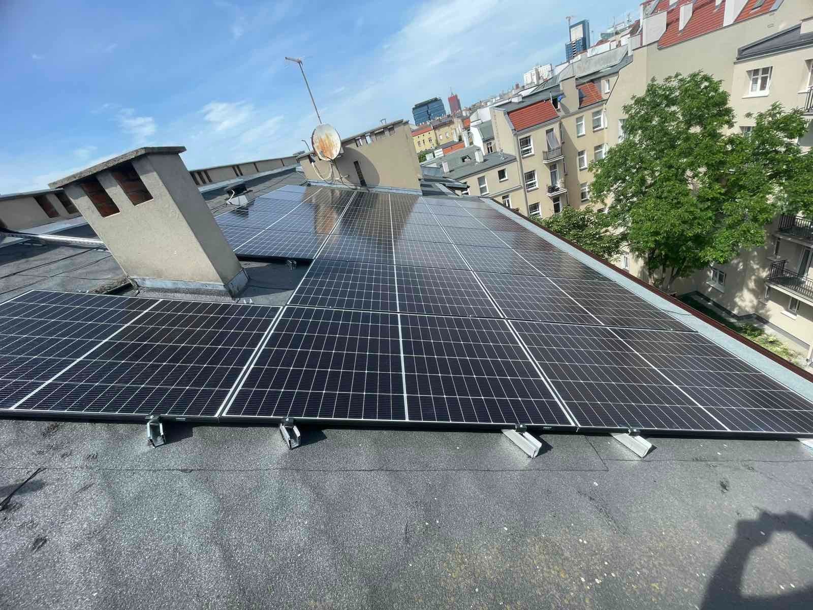 Panele słoneczne na dachu budynku w miejskiej scenerii, widoczne mocowania i komin. W tle zabudowa miejska i błękitne niebo.