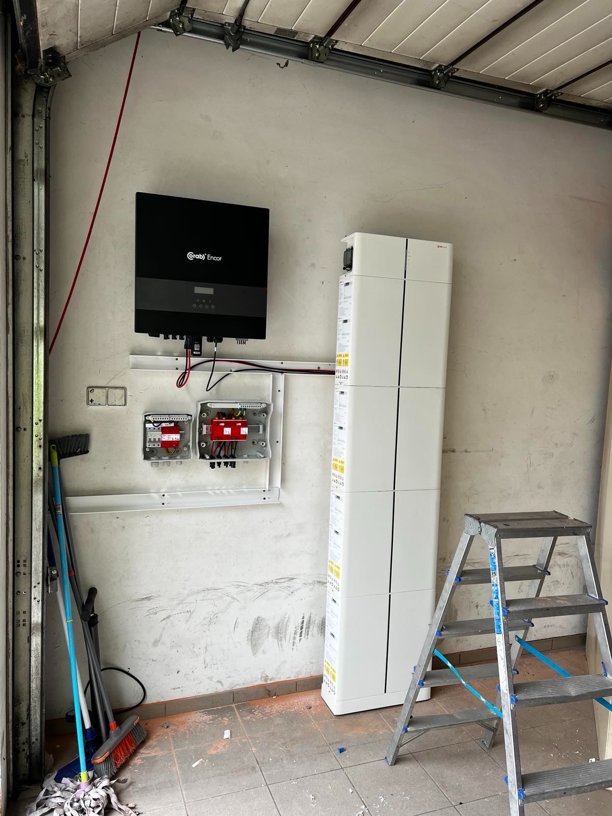 Montaż magazynu energii Encor i inwertera na ścianie garażu. Widoczne kable, skrzynki elektryczne i drabina. Podłoga z drobnymi zabrudzeniami.