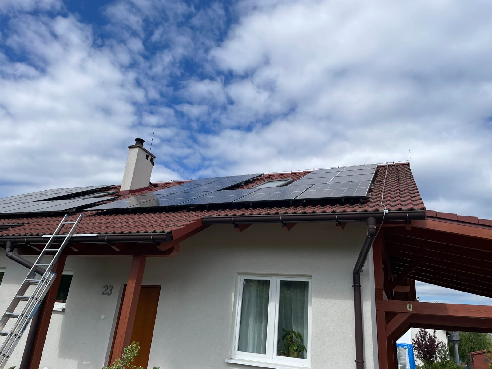 Dom jednorodzinny z panelami słonecznymi na dachu z czerwonej dachówki. Aluminiowa drabina oparta o ścianę. Błękitne niebo z chmurami w tle.