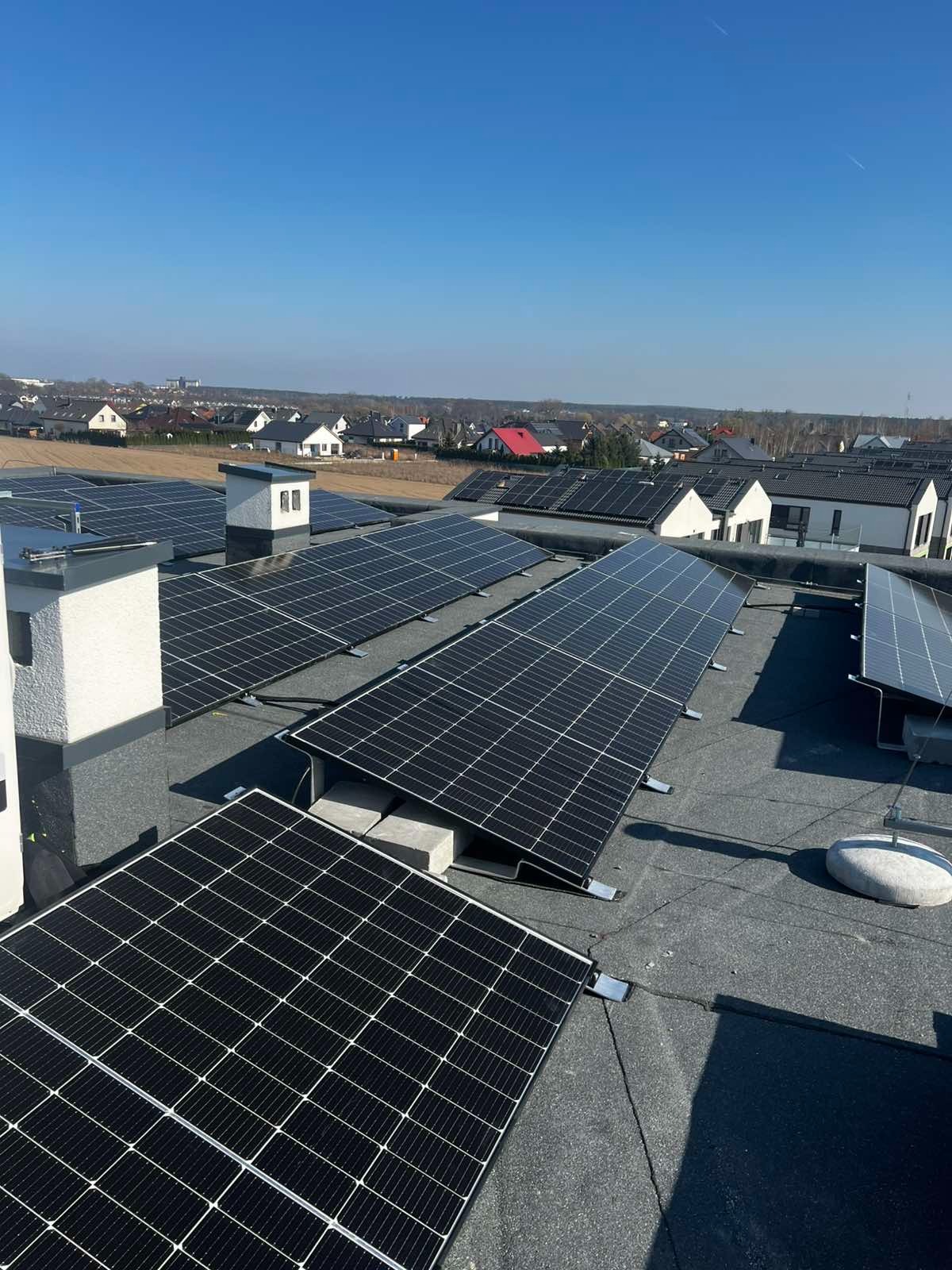 Instalacja fotowoltaiczna na dachu budynku z kominami, widok z góry na tle zabudowy jednorodzinnej i błękitnego nieba w słoneczny dzień. Panele zamontowane na betonowych podstawach.
