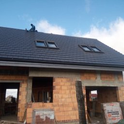 Przemysław Rejek Dekarstwo - Nowy dach z dachówką ceramiczną i oknami połaciowymi na budynku w trakcie budowy. Widoczny dekarz na dachu i materiały budowlane. Ujęcie z dołu.